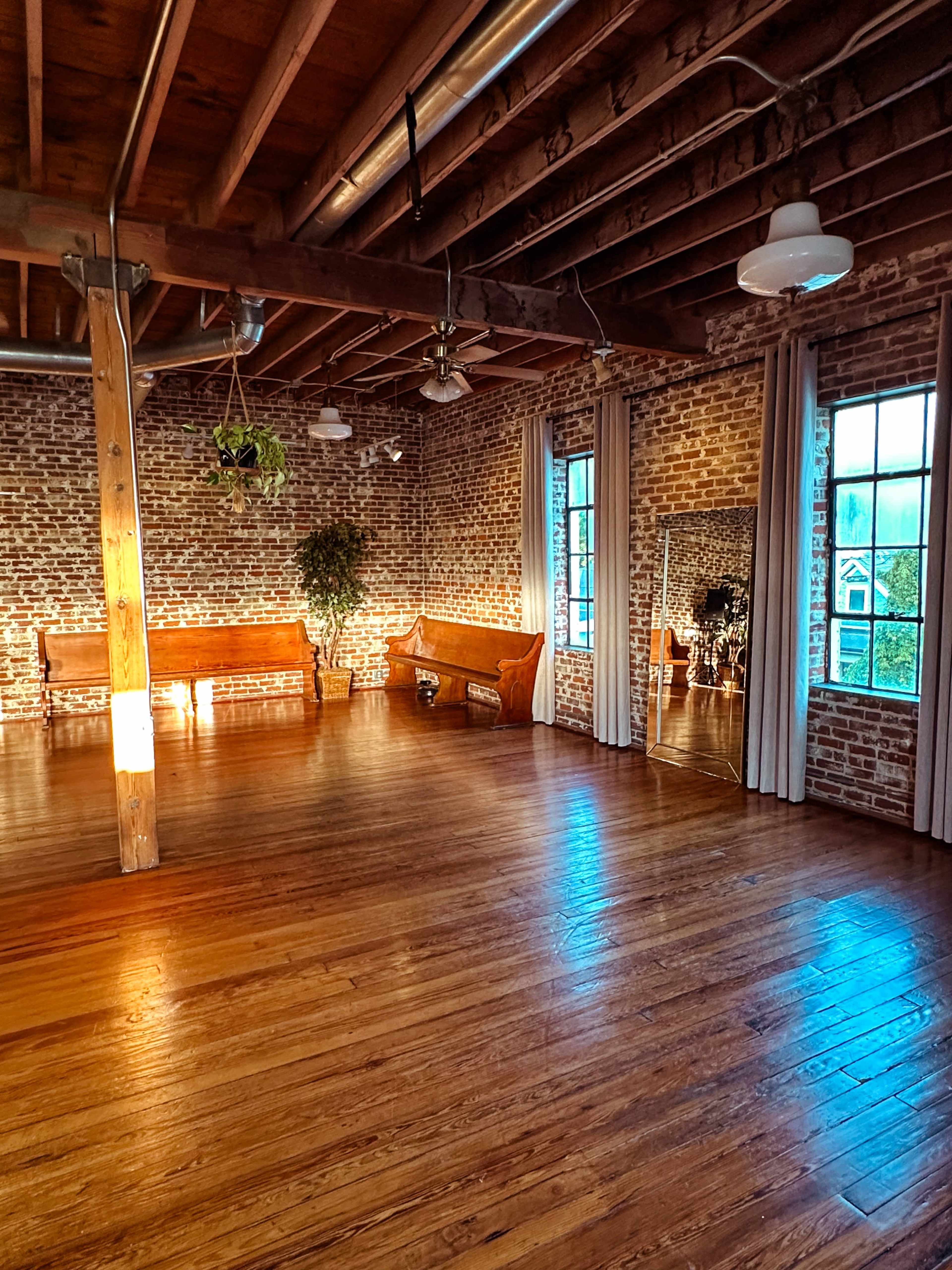 The image shows a spacious room with exposed brick walls, wooden floors, and large windows, featuring two benches and a mirror along one wall.