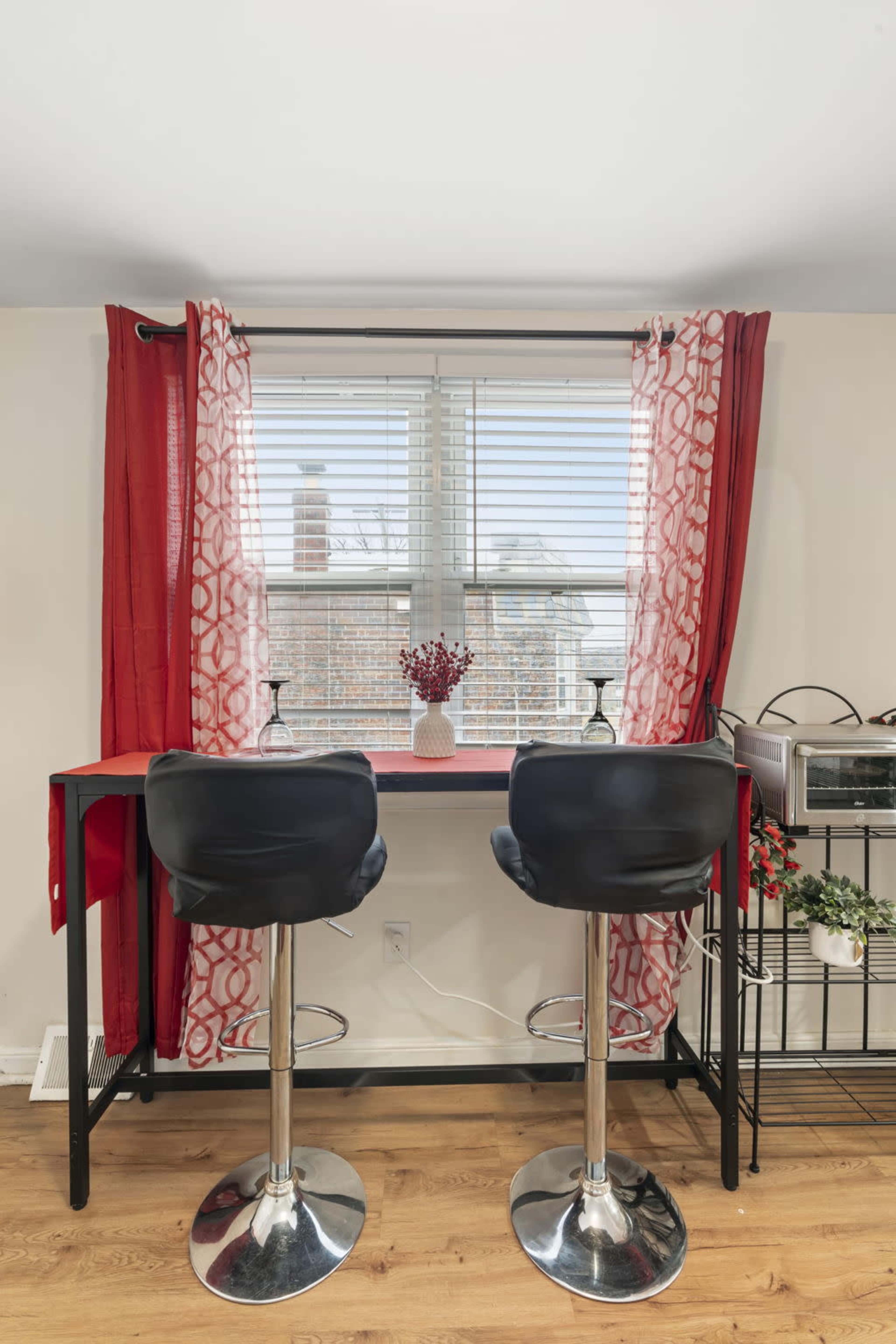 A small desk with two black bar stools is positioned by a window covered with red curtains, while a vase of flowers sits on the desk.