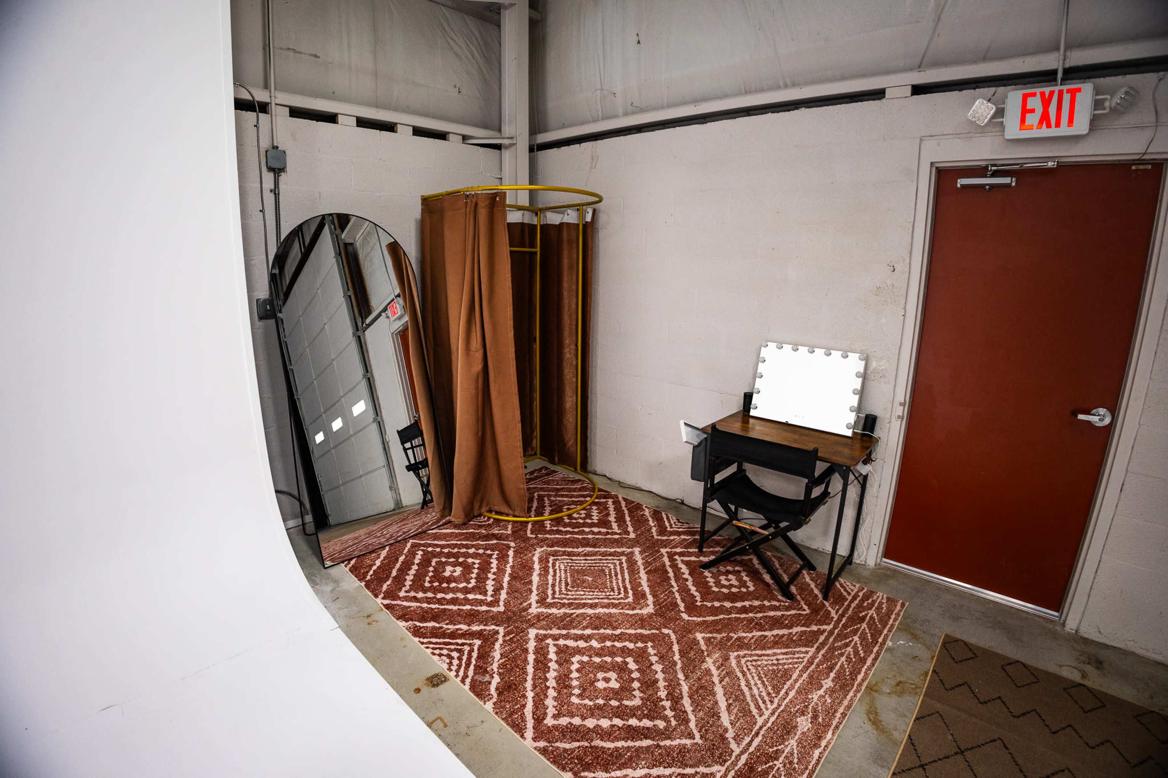 The image shows a small, minimalistic room featuring a dressing area with a mirror, a brown curtain, a chair, and a lighted vanity table on a patterned rug.