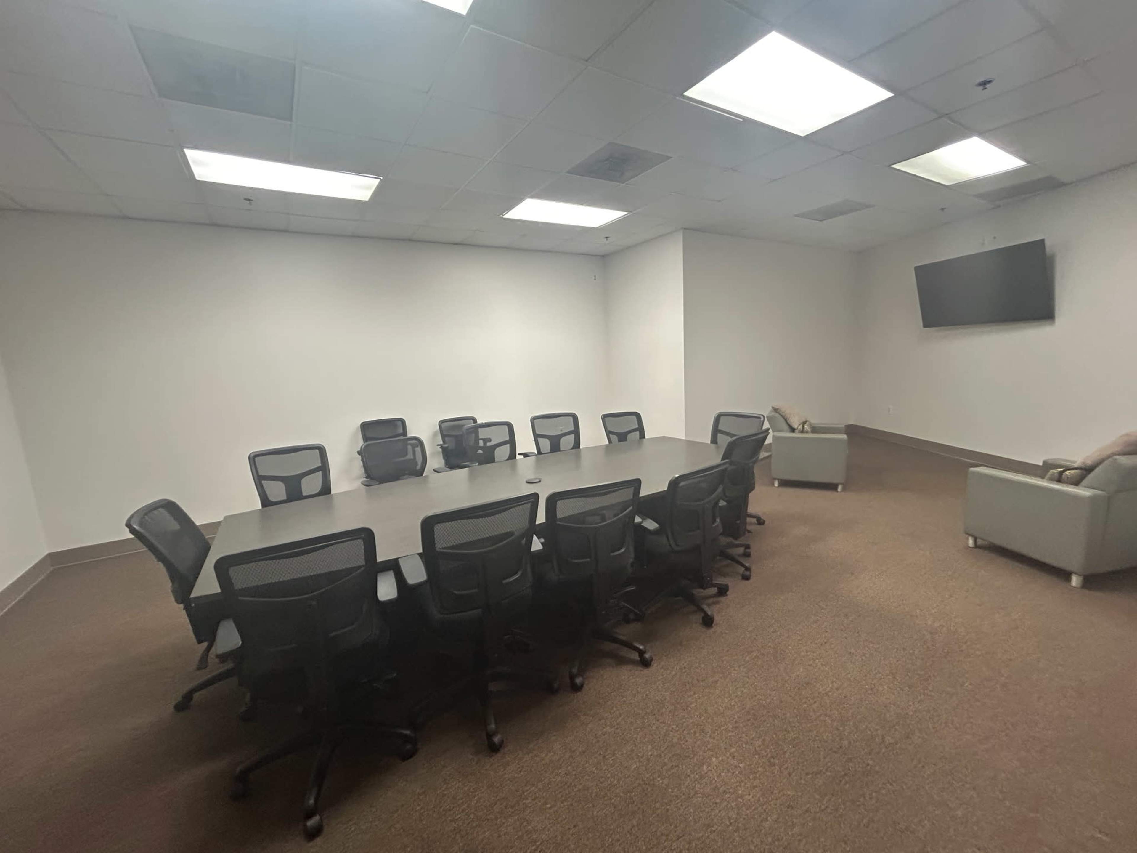 A large conference room features a rectangular table surrounded by several office chairs, with a couch visible in the corner and a television mounted on the wall.