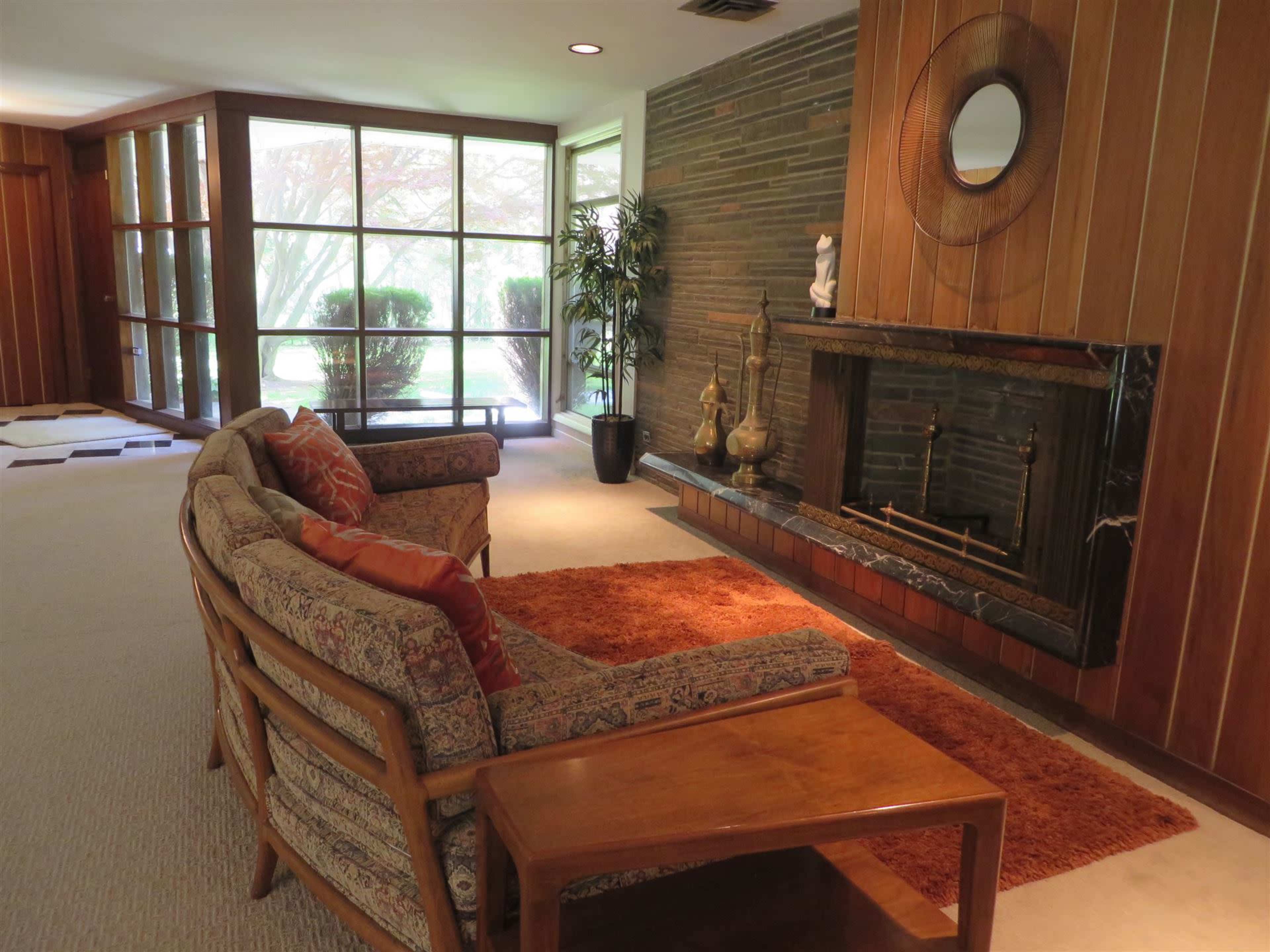 The image shows a living room with a stone fireplace, a patterned sofa set, and a large window overlooking a garden.