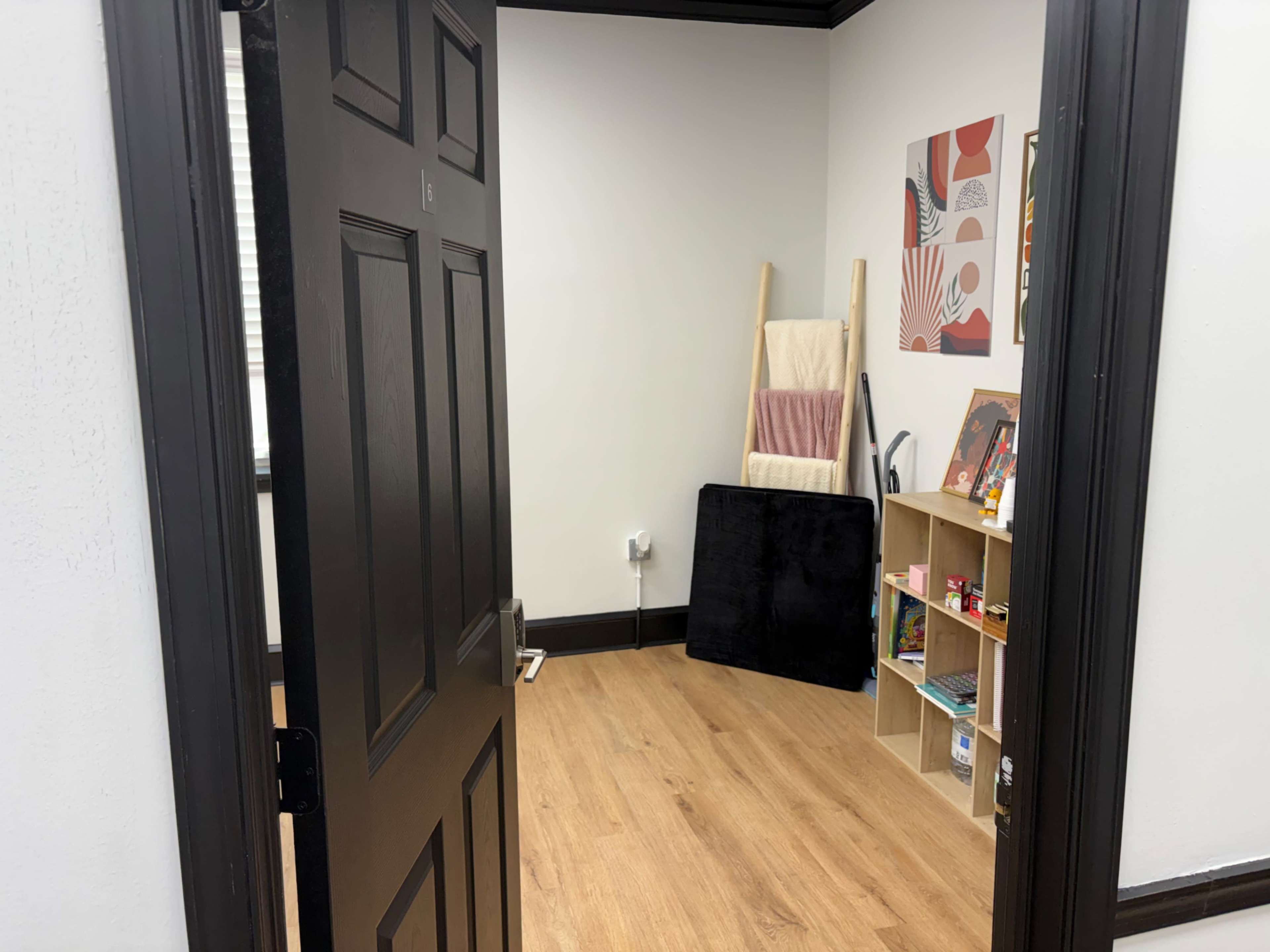 The image shows a doorway leading into a small room with wooden flooring, a bookshelf filled with colorful items, and a minimalist decor featuring an easel and a black fabric draped on the floor.