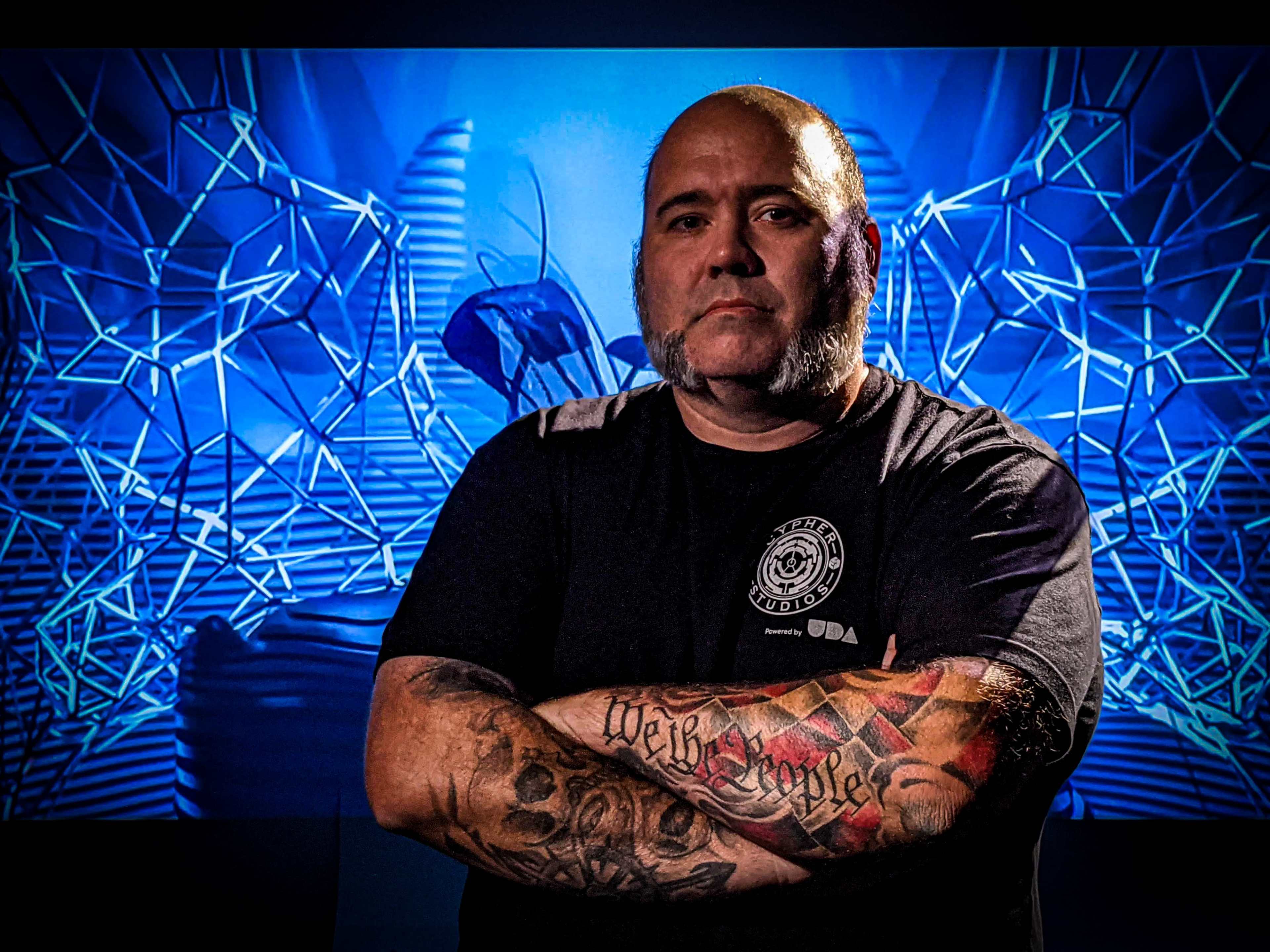 A man with tattoos on his arms stands cross-armed in front of a digitally created background featuring abstract shapes.