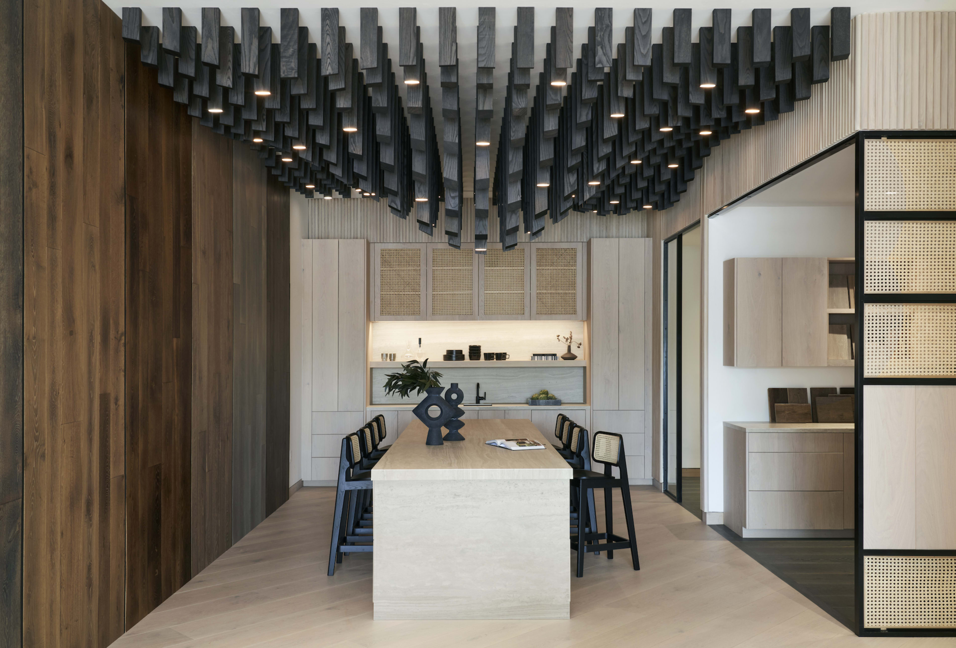 The image shows a modern dining area with a large light-colored table surrounded by black chairs, featuring a unique wooden ceiling design overhead.