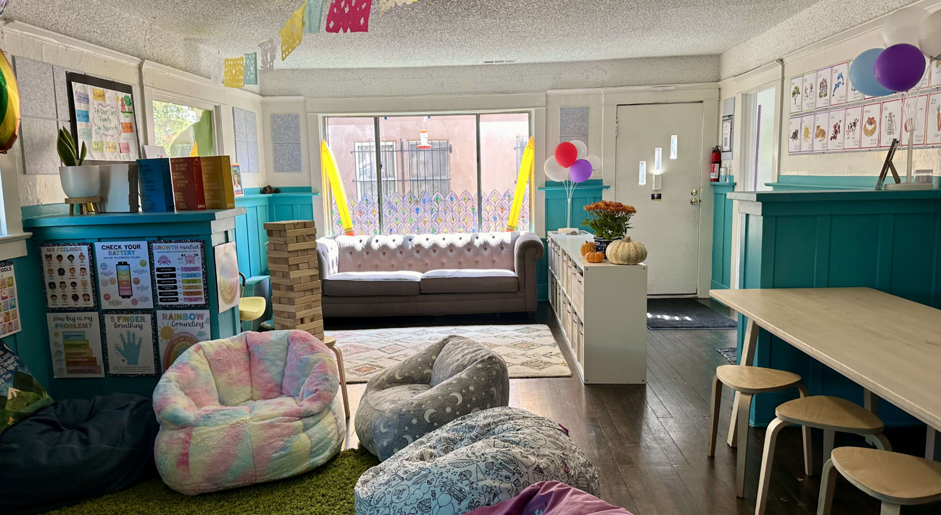 The room features colorful bean bags, a couch, a table, and decorated walls with various educational posters and balloons.