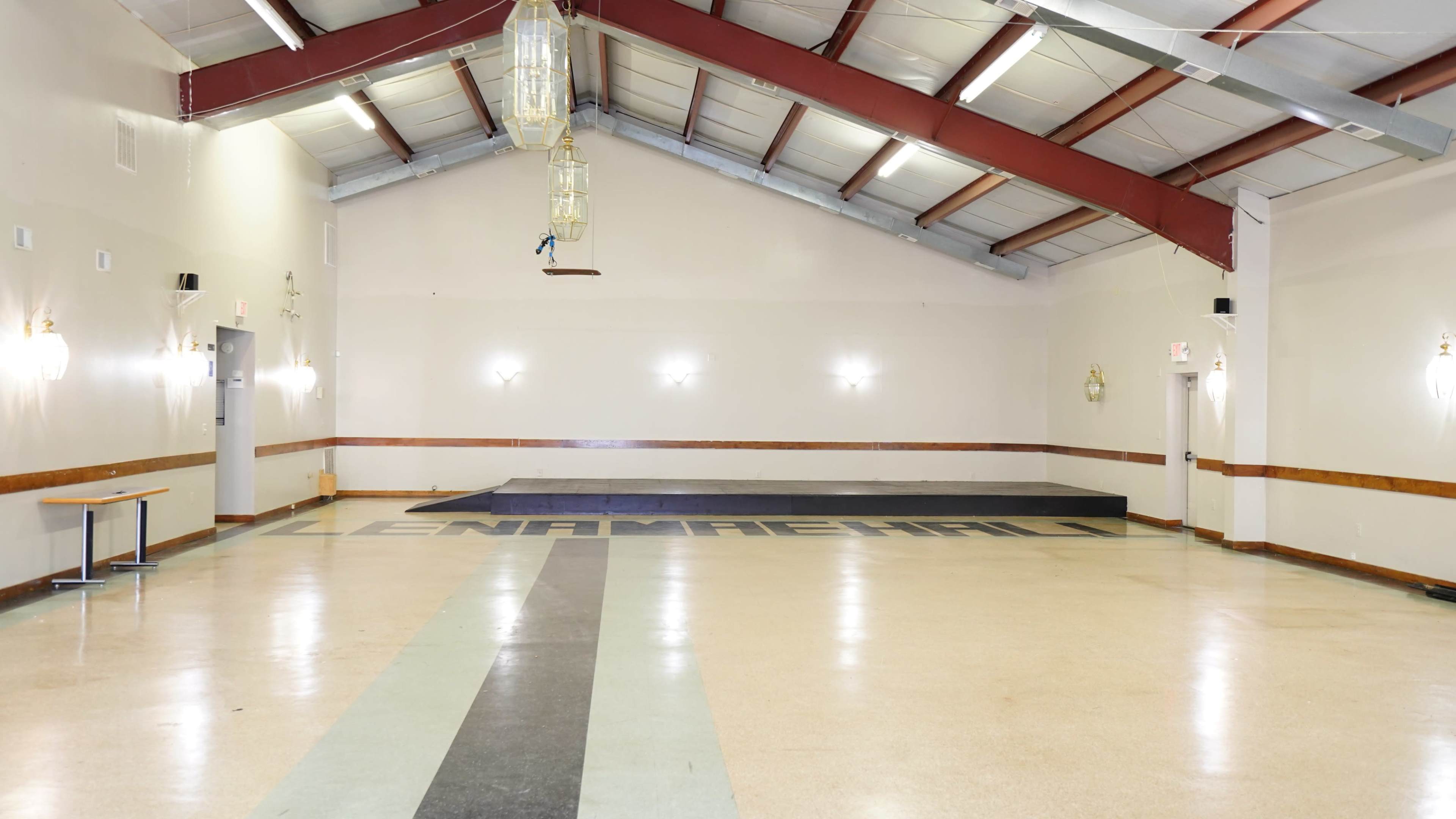 An empty hall with a wooden platform at one end and "LENAMAREHALL" painted on the floor.