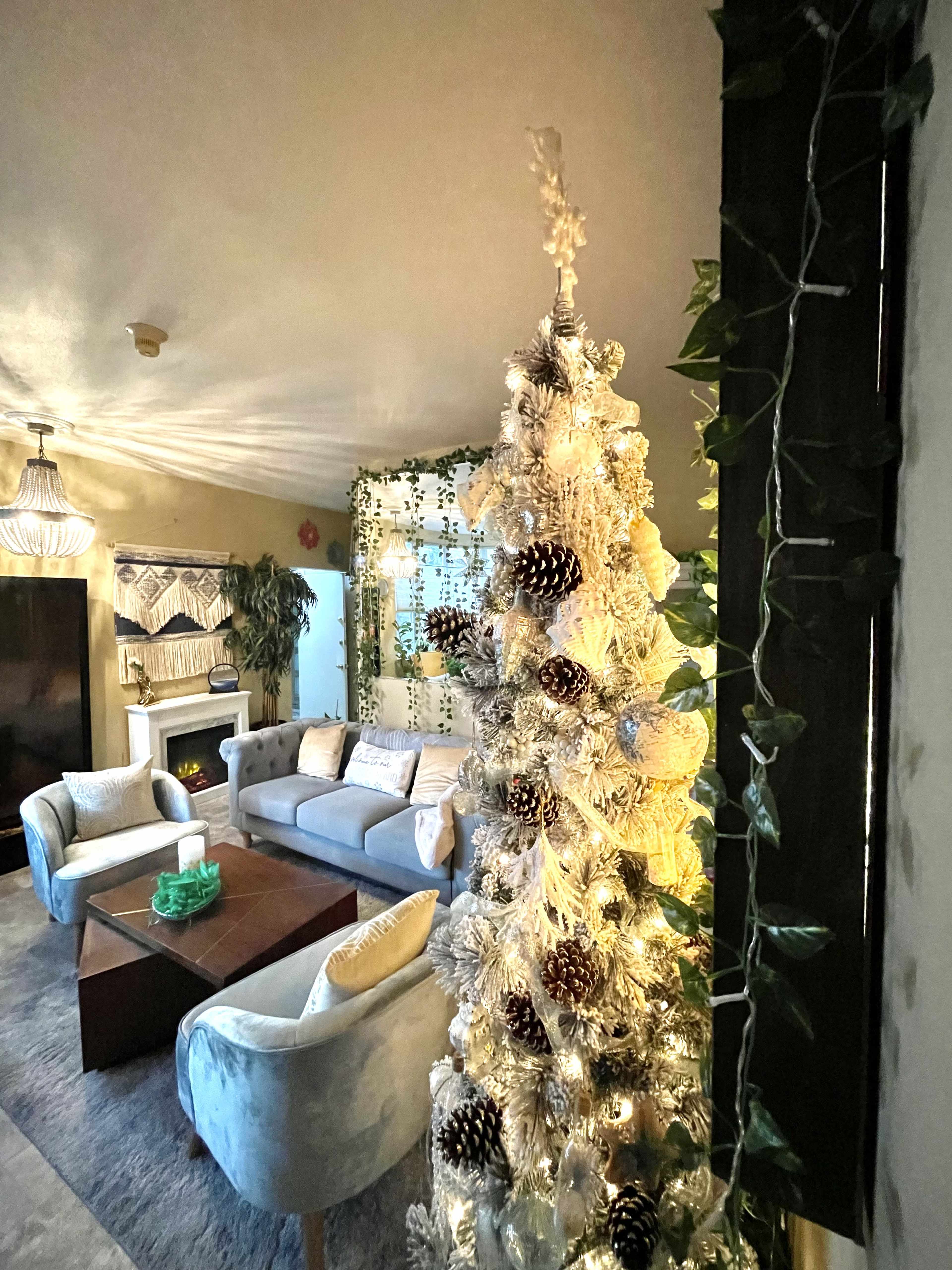 A living room decorated for the holidays, featuring a white Christmas tree adorned with pine cones and illuminated by lights, along with a gray sofa and a coffee table in the center.