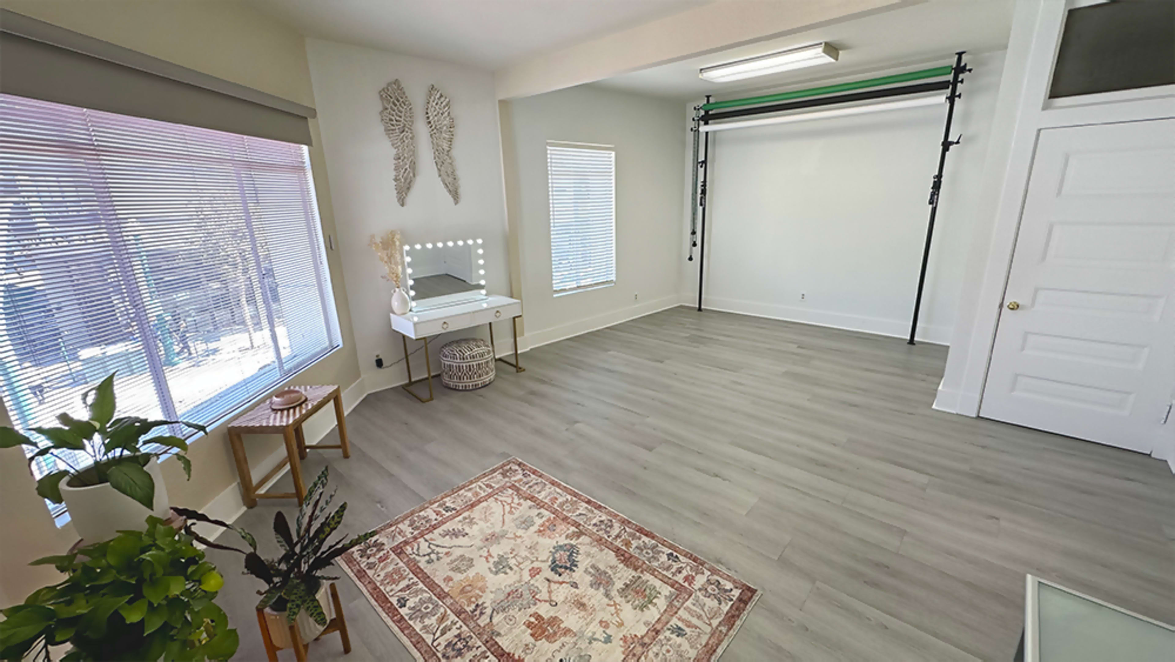 A well-lit, empty room with large windows, a small table, a decorative rug, and a mirror.