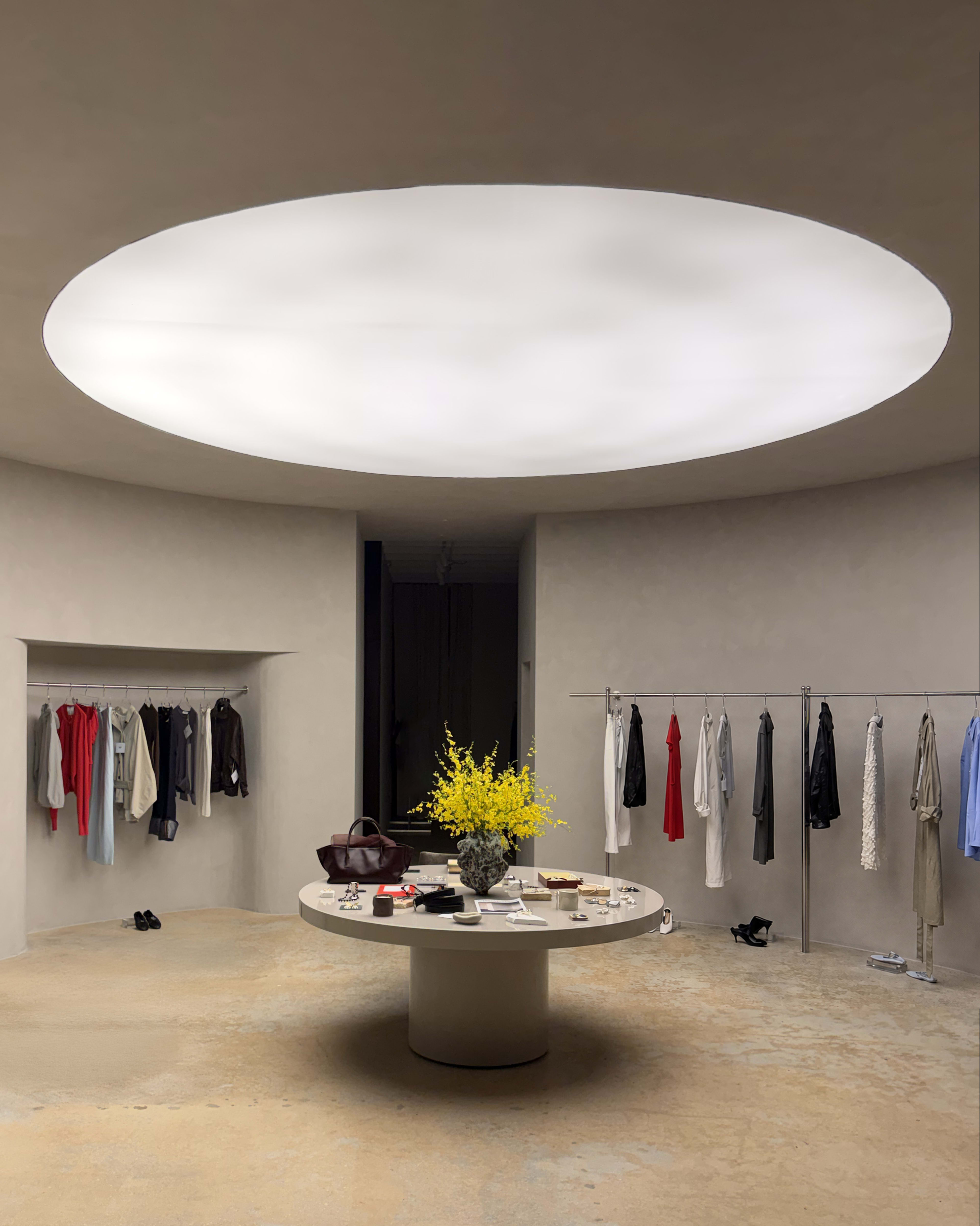 The image shows a modern, minimalist fashion boutique with a circular table in the center and clothing hung on racks around the walls, illuminated by a large circular skylight.
