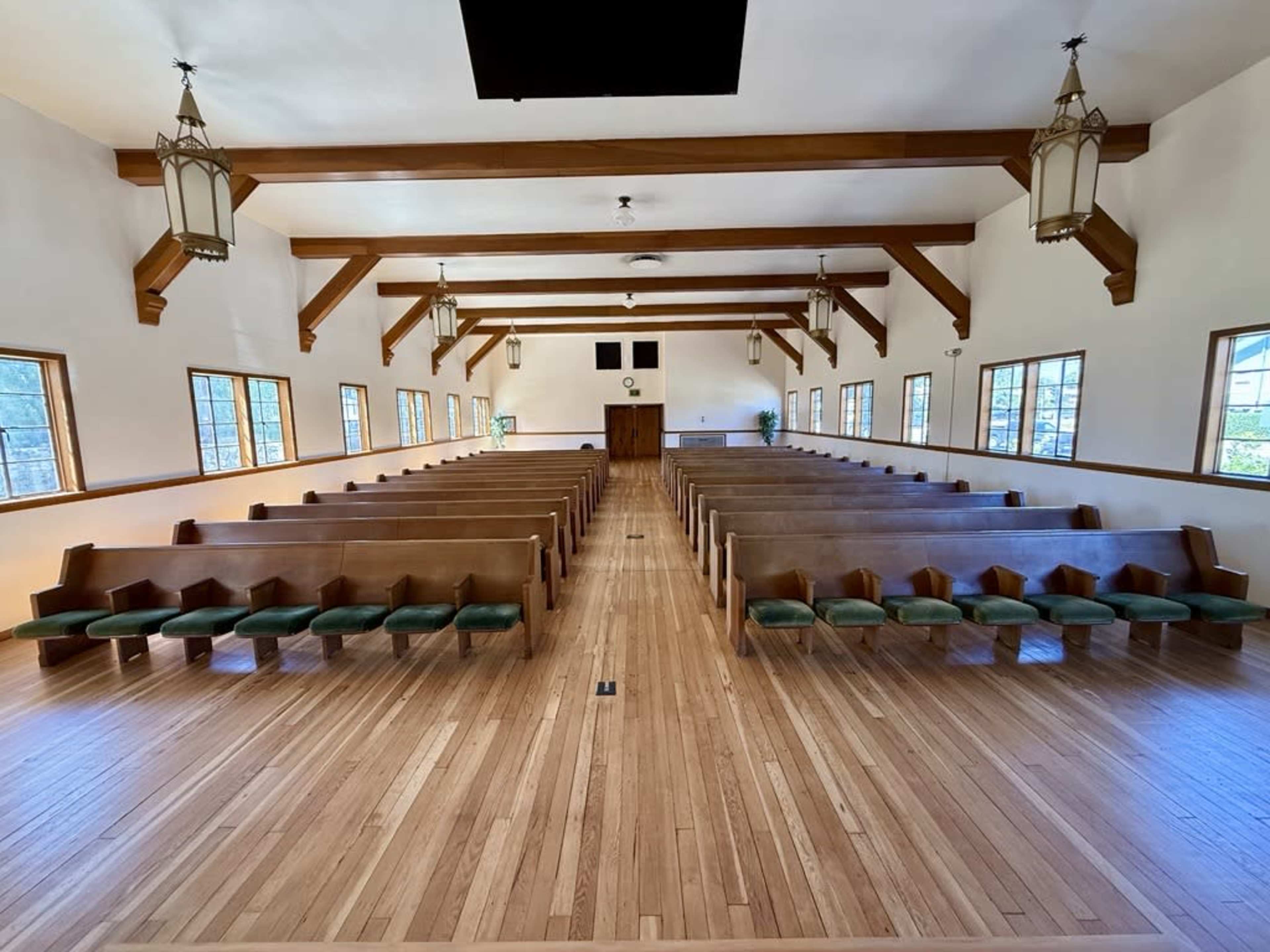 The image shows a spacious interior of a church with wooden pews arranged in neat rows on a polished wooden floor under wooden beams and lanterns.