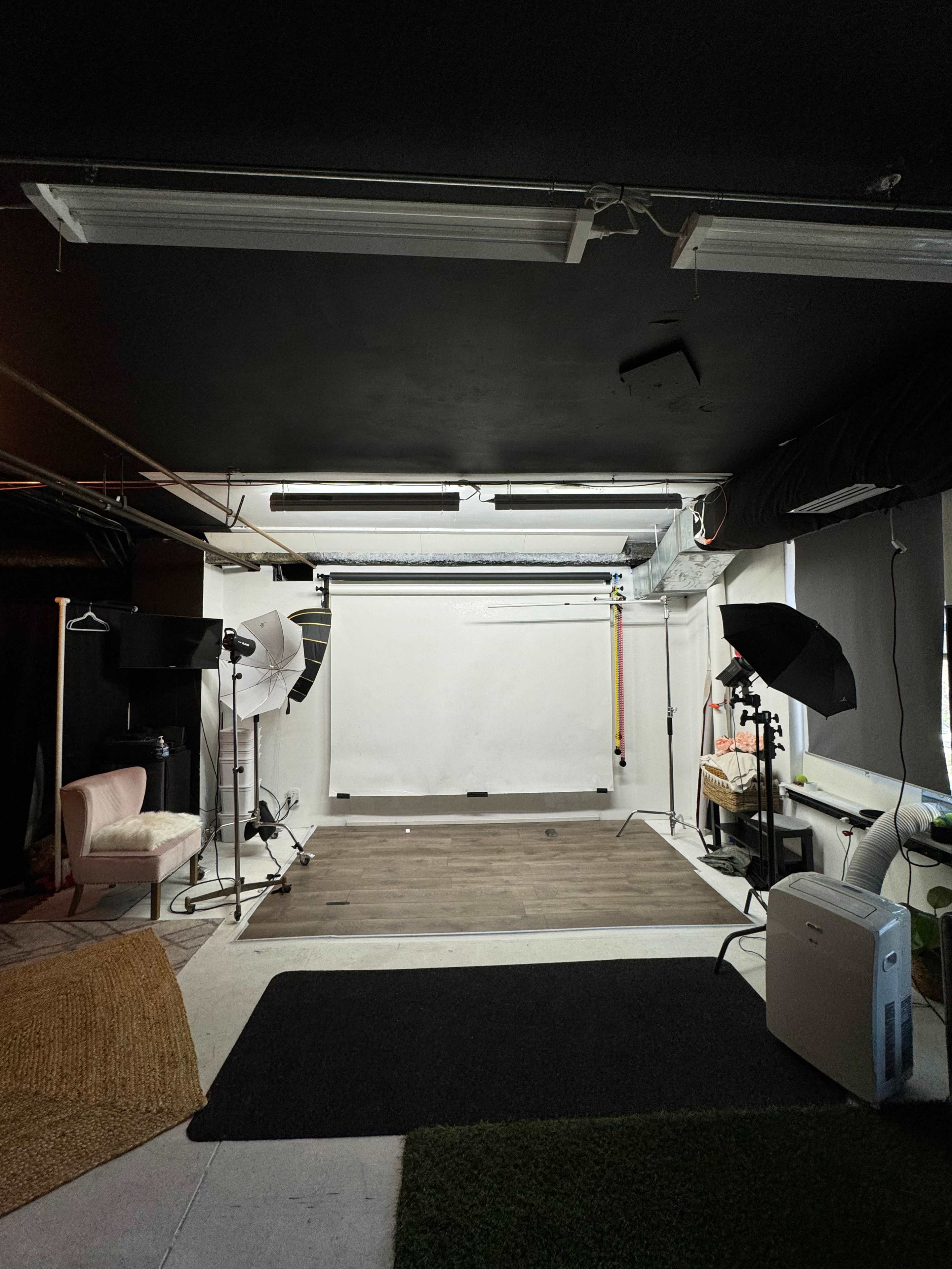 The image shows a photography studio featuring a blank backdrop, lighting equipment, and a combination of hard floor and carpeted areas.