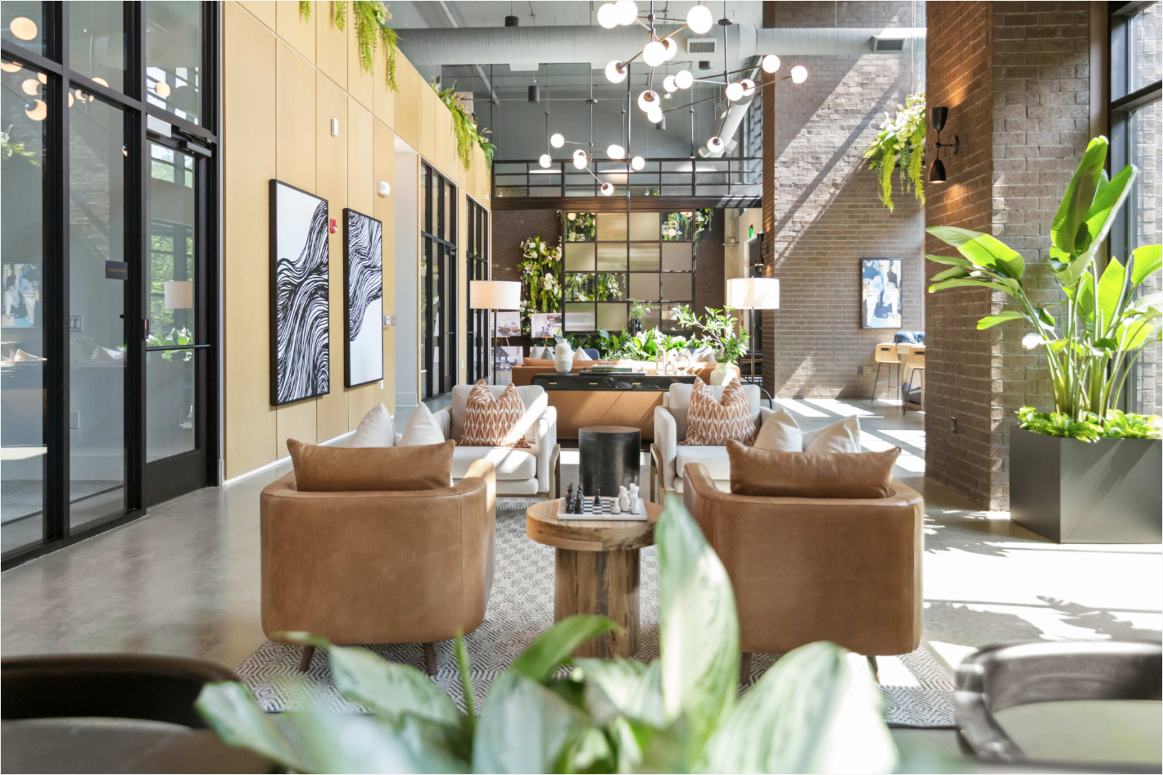 The image shows a modern lounge area featuring comfortable seating, decorative plants, and artwork against a backdrop of large windows and stylish architecture.