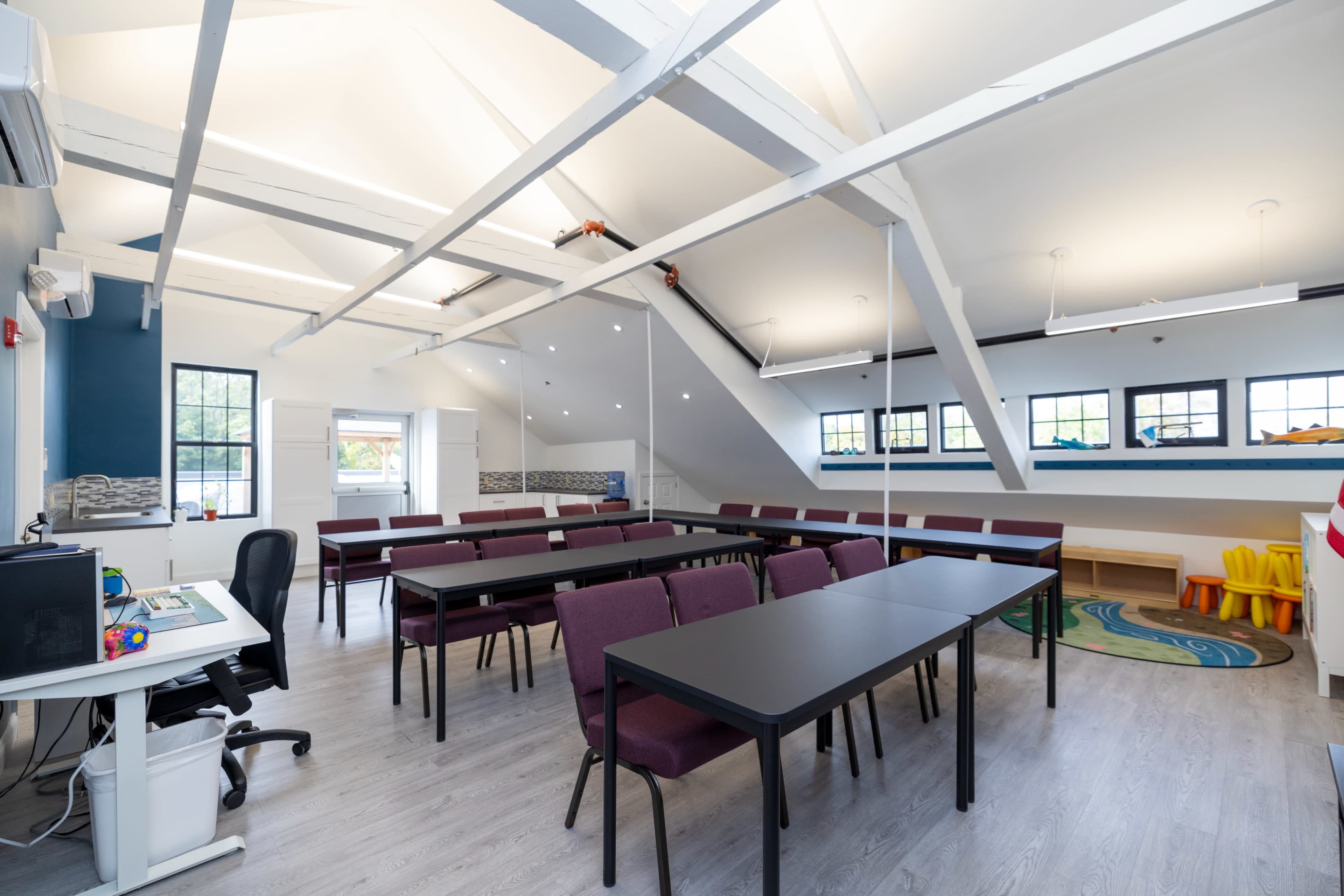 A spacious classroom with several black tables and purple chairs arranged in rows under a sloped ceiling.
