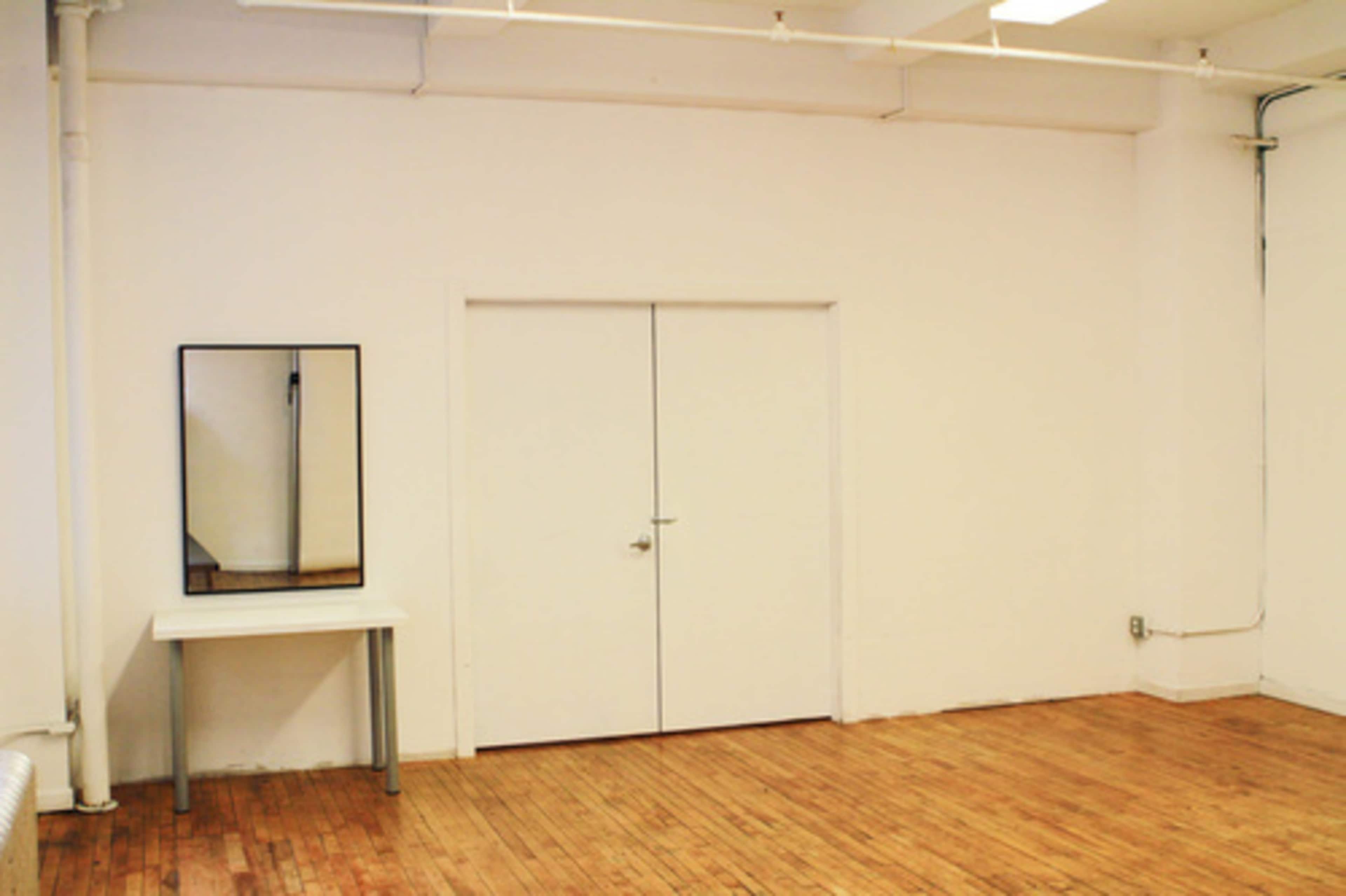 A minimalist room with wooden flooring, a white wall, a large mirror above a table, and double doors.