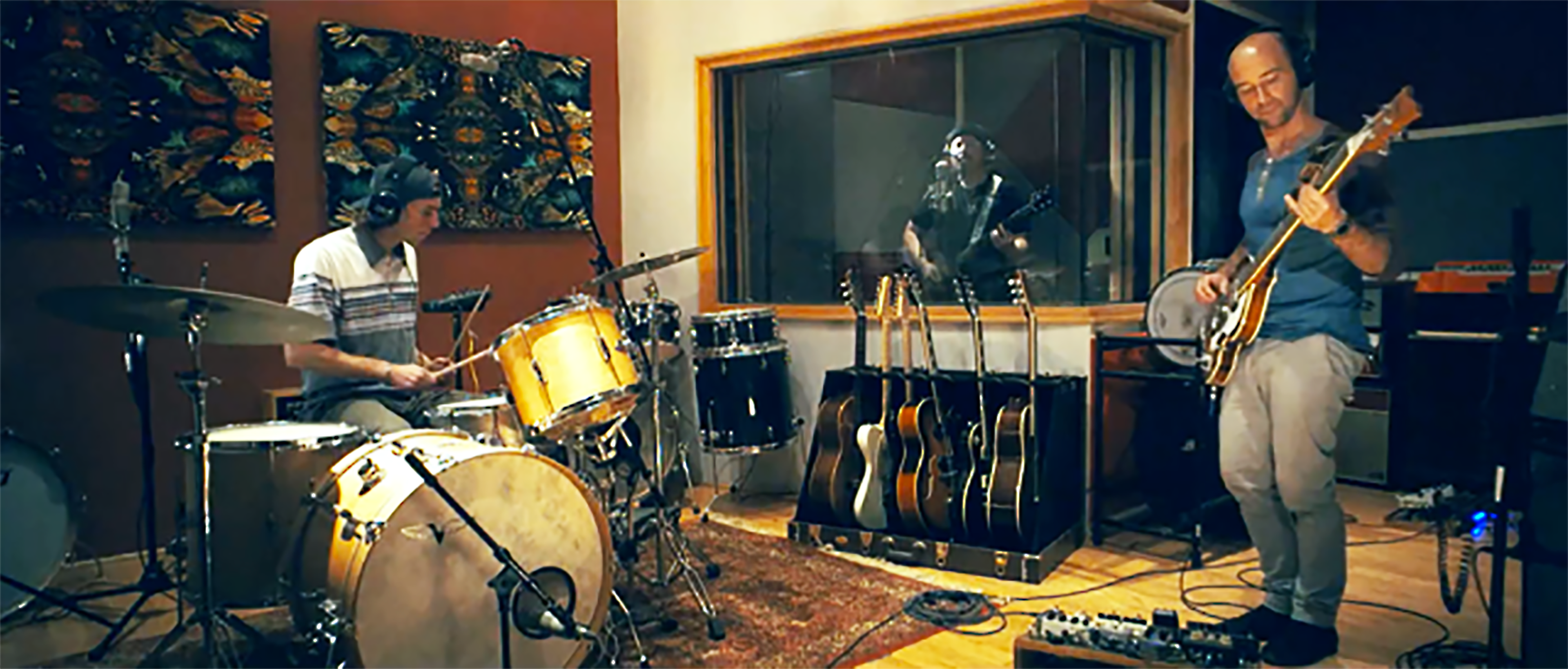Two musicians are playing instruments in a recording studio, with one on drums and the other on guitar, while a vocalist is visible behind a glass panel.