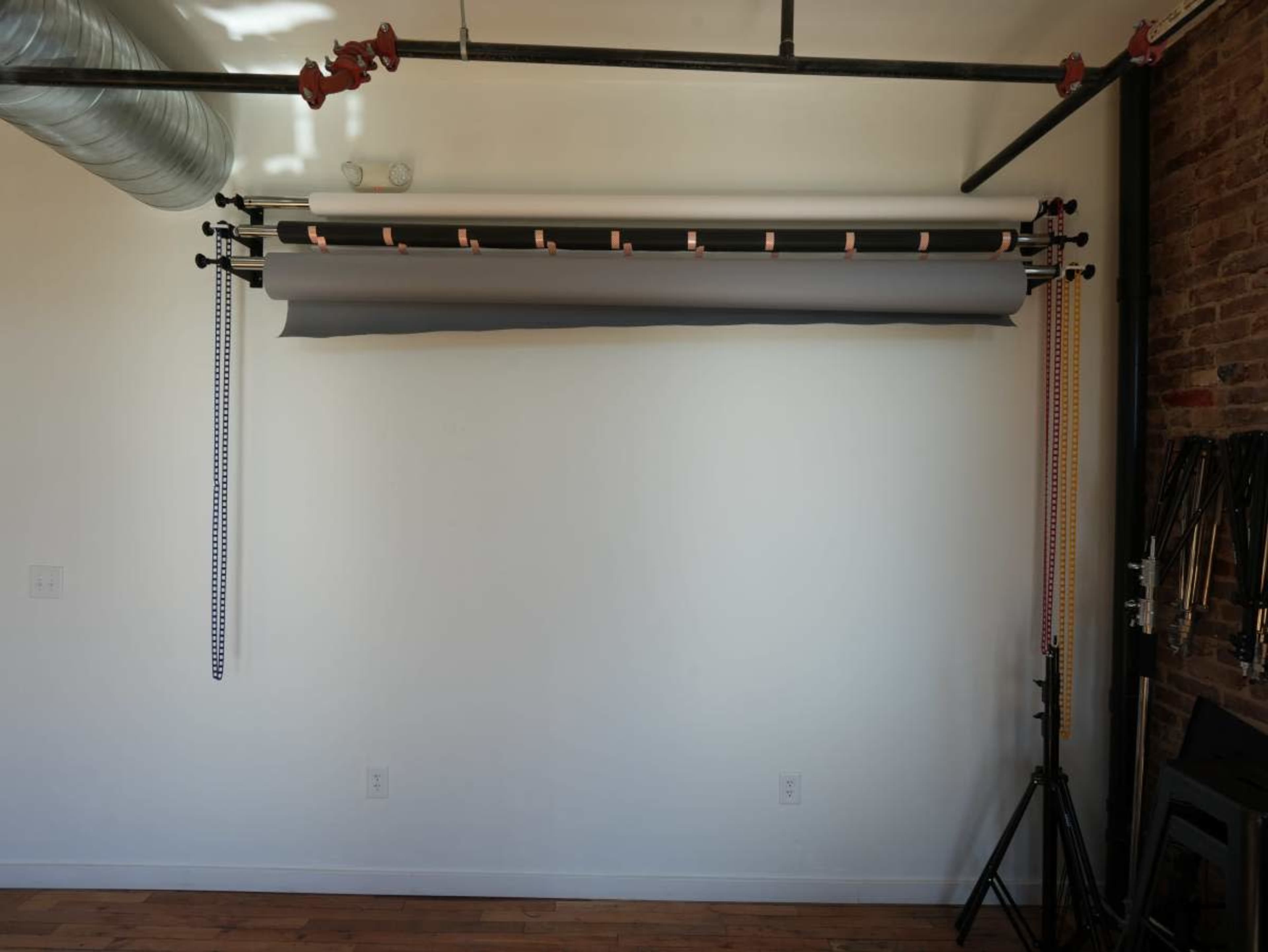 The image shows a photography backdrop setup with a gray roll of paper mounted on a horizontal bar against a white wall in a room with wooden flooring.
