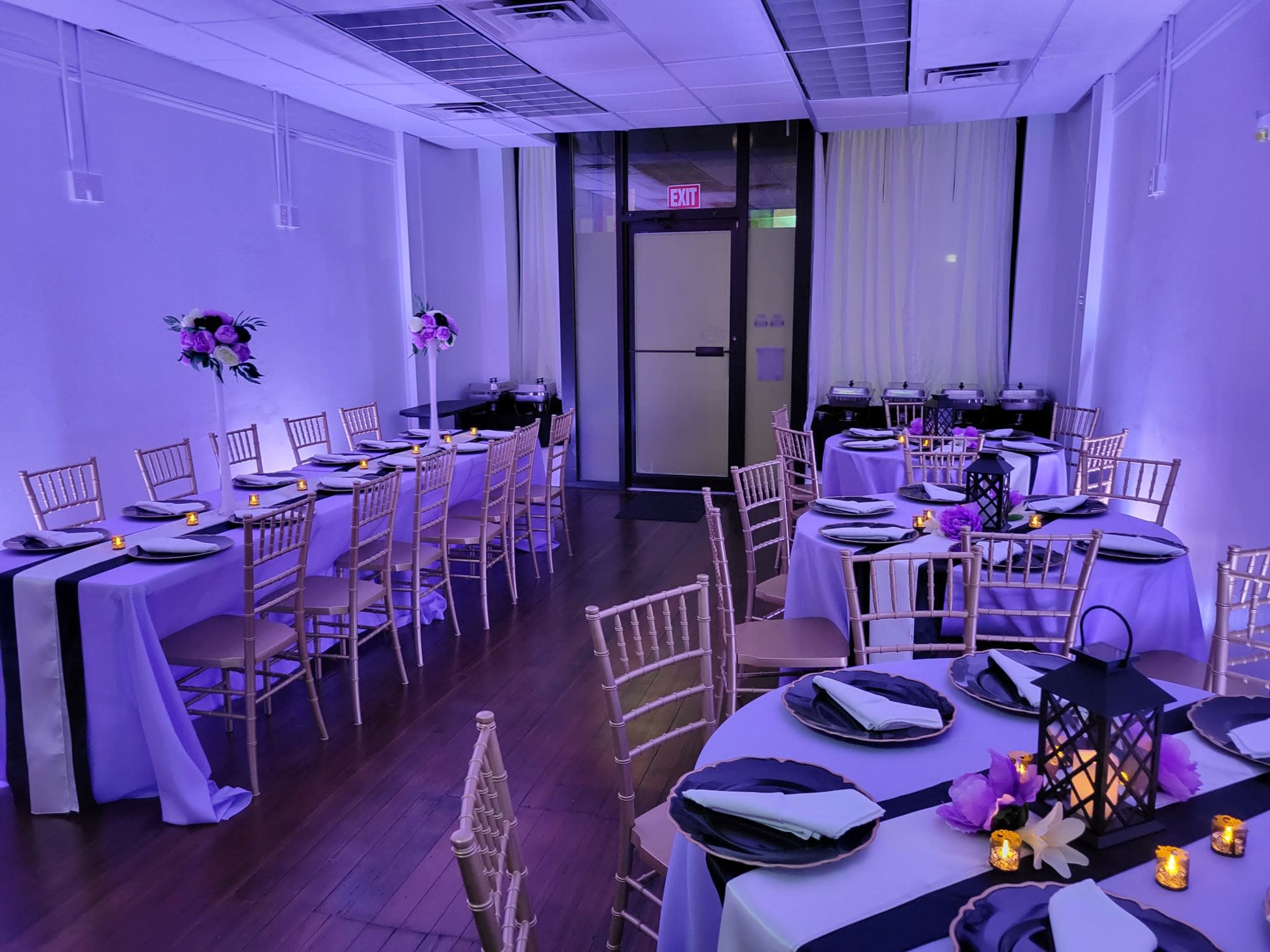 The image shows a beautifully arranged banquet hall with several tables set for dining, featuring purple and black tablecloths, candles, and floral centerpieces.
