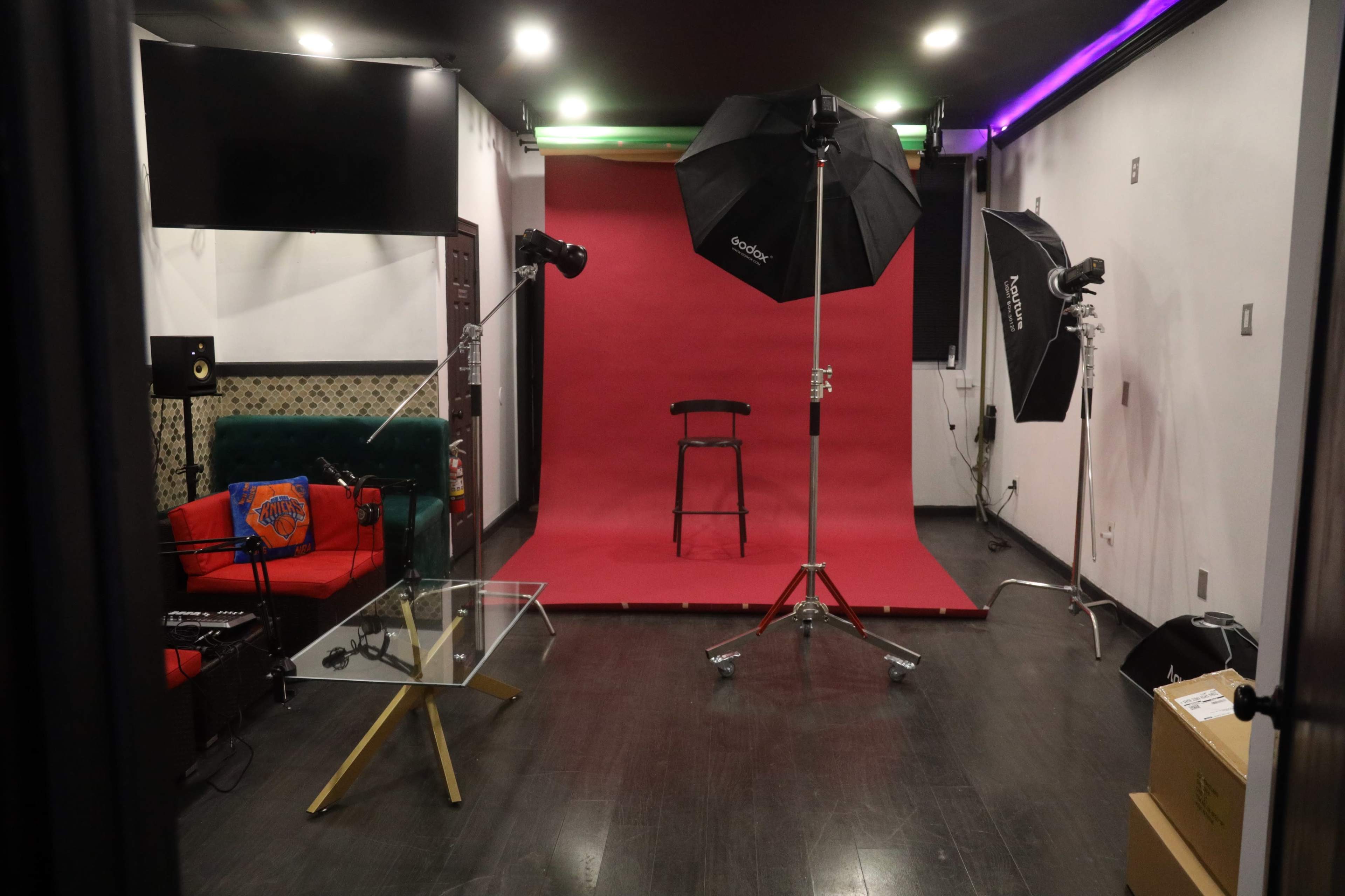 A photography studio is set up with a red backdrop, a black chair, and lighting equipment, surrounded by a green couch and a glass table.
