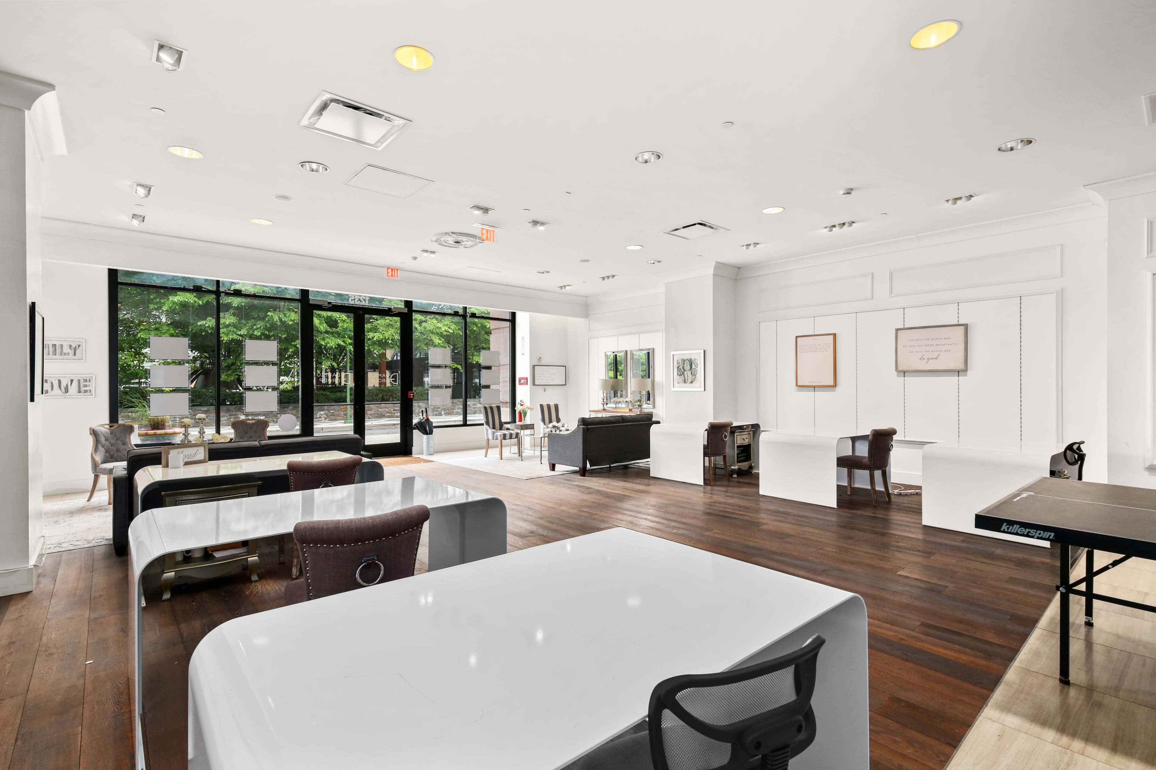 A spacious, modern office interior with large windows, white walls, and wooden floors, featuring desks, chairs, and a ping pong table.