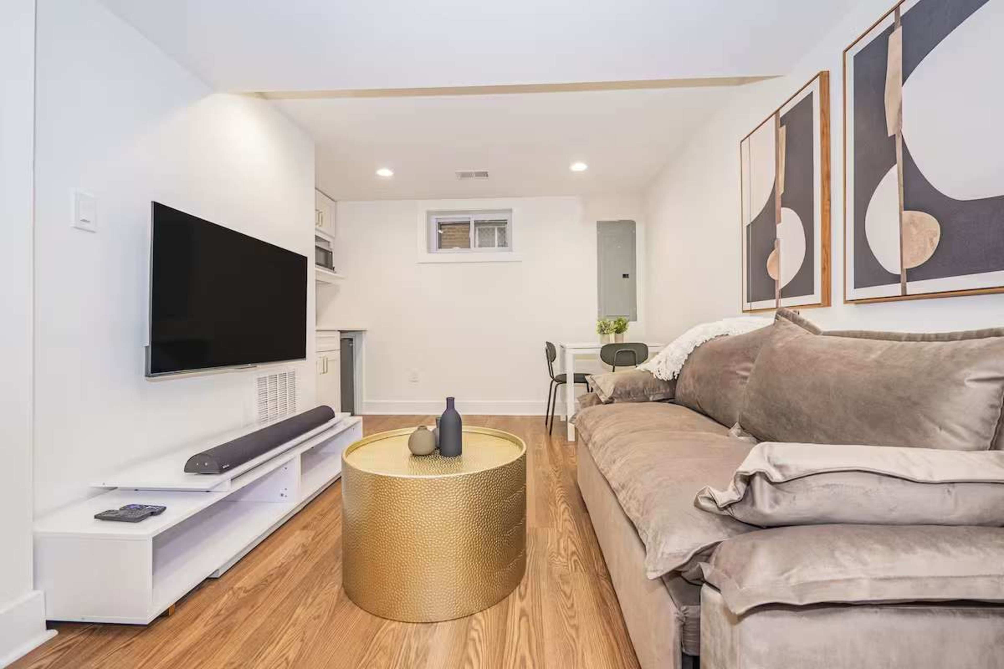 The image shows a cozy living area featuring a large sofa, a round coffee table, a wall-mounted television, and abstract artwork on the walls.