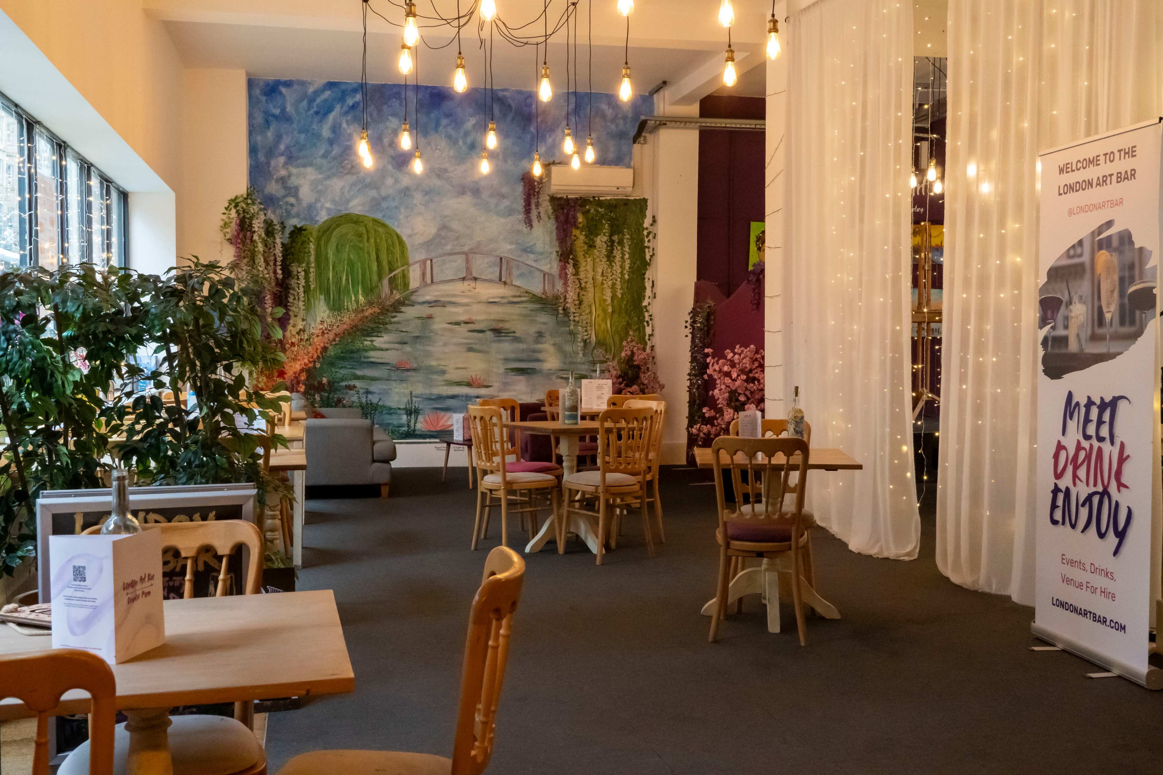 The image shows a cozy interior of a bar with wooden tables, chairs, a large mural depicting a landscape, and decorative lighting.