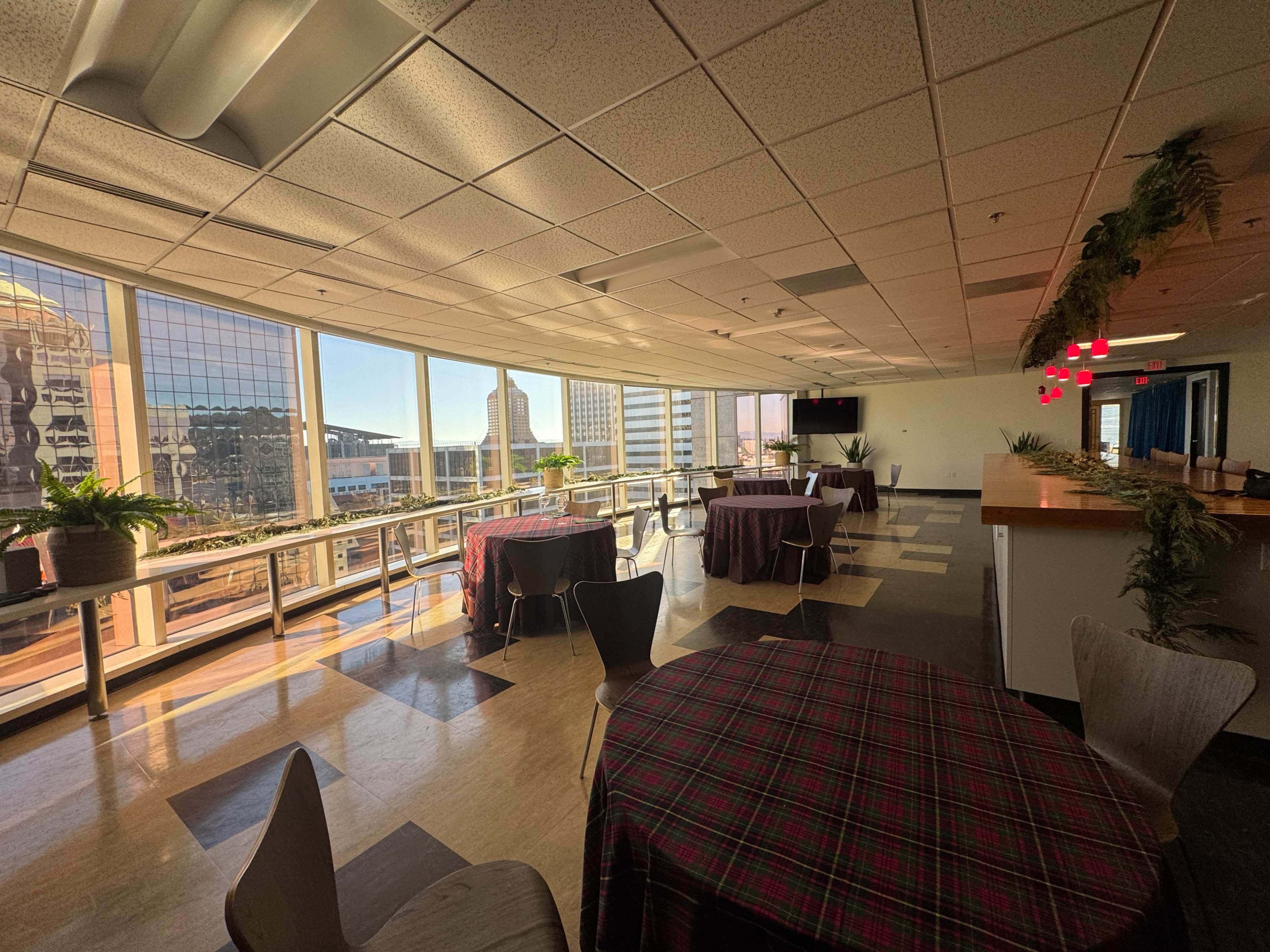 The image shows a spacious, well-lit room with tables covered in checkered cloths, large windows revealing a cityscape, and a bar area in the background.