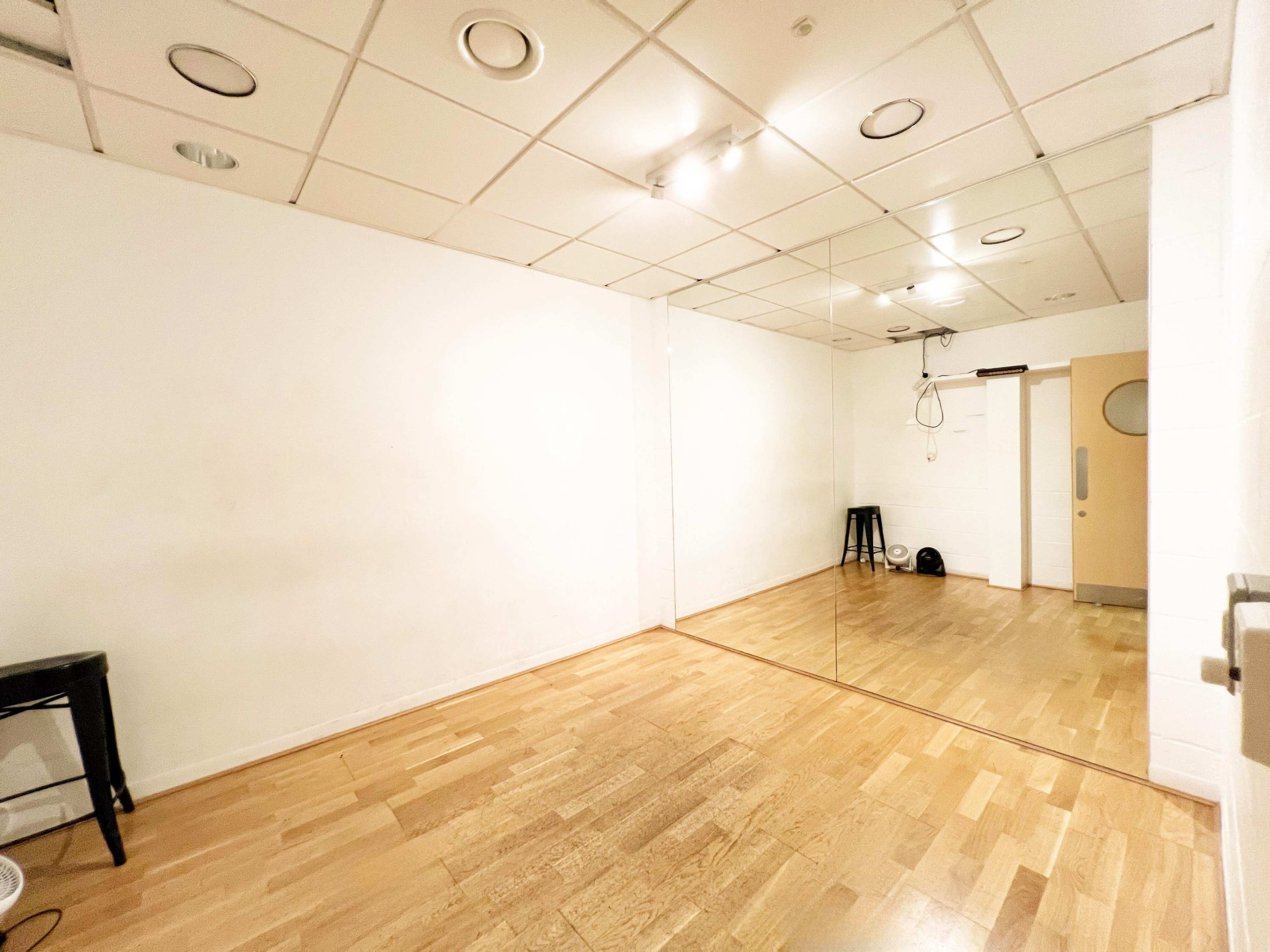 The image shows a spacious, empty room with wooden flooring and a large mirror on one wall.