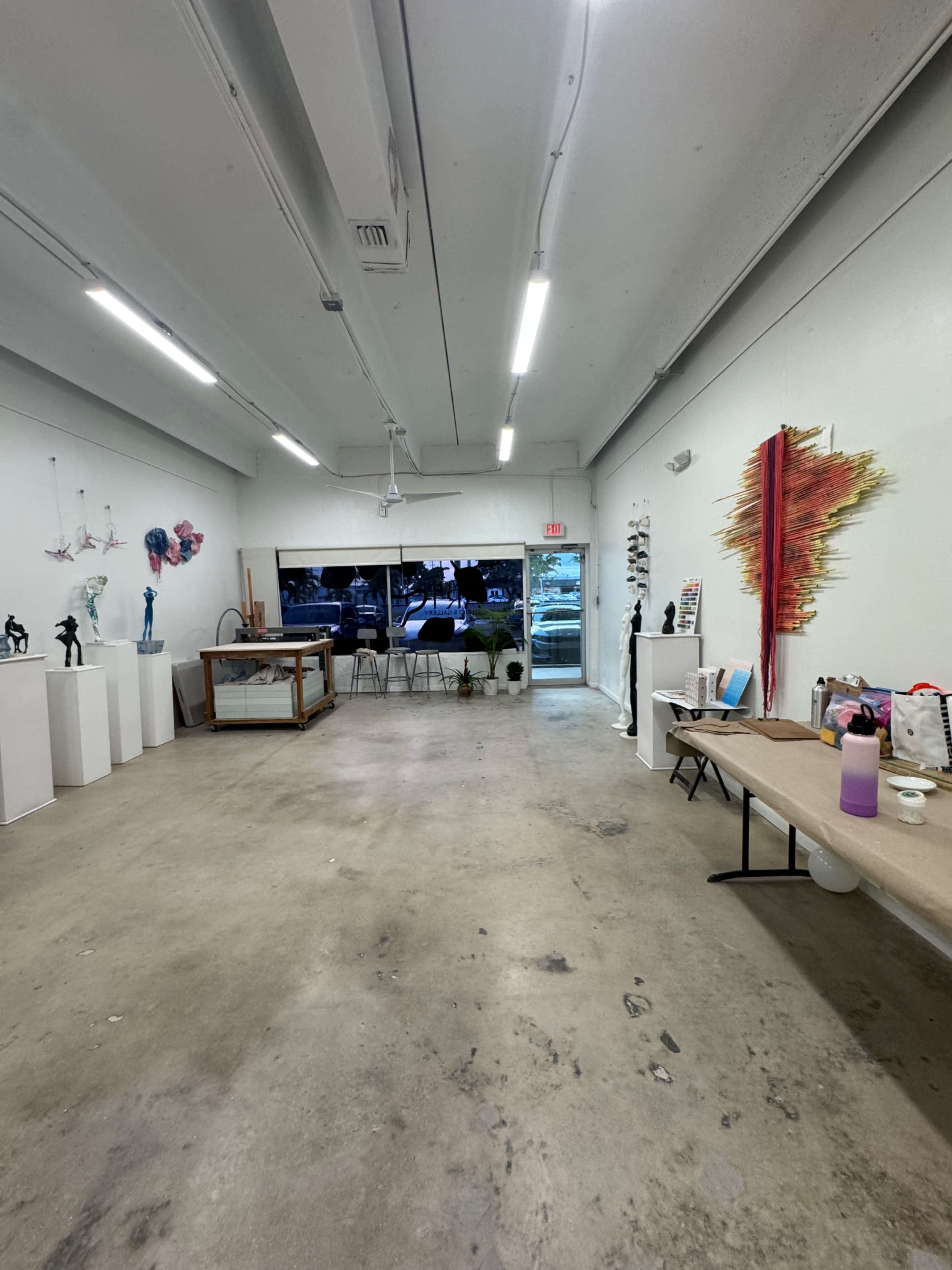 The image shows an empty art gallery space featuring sculpture displays on white pedestals, a table with art supplies, and a large window revealing an outdoor view.