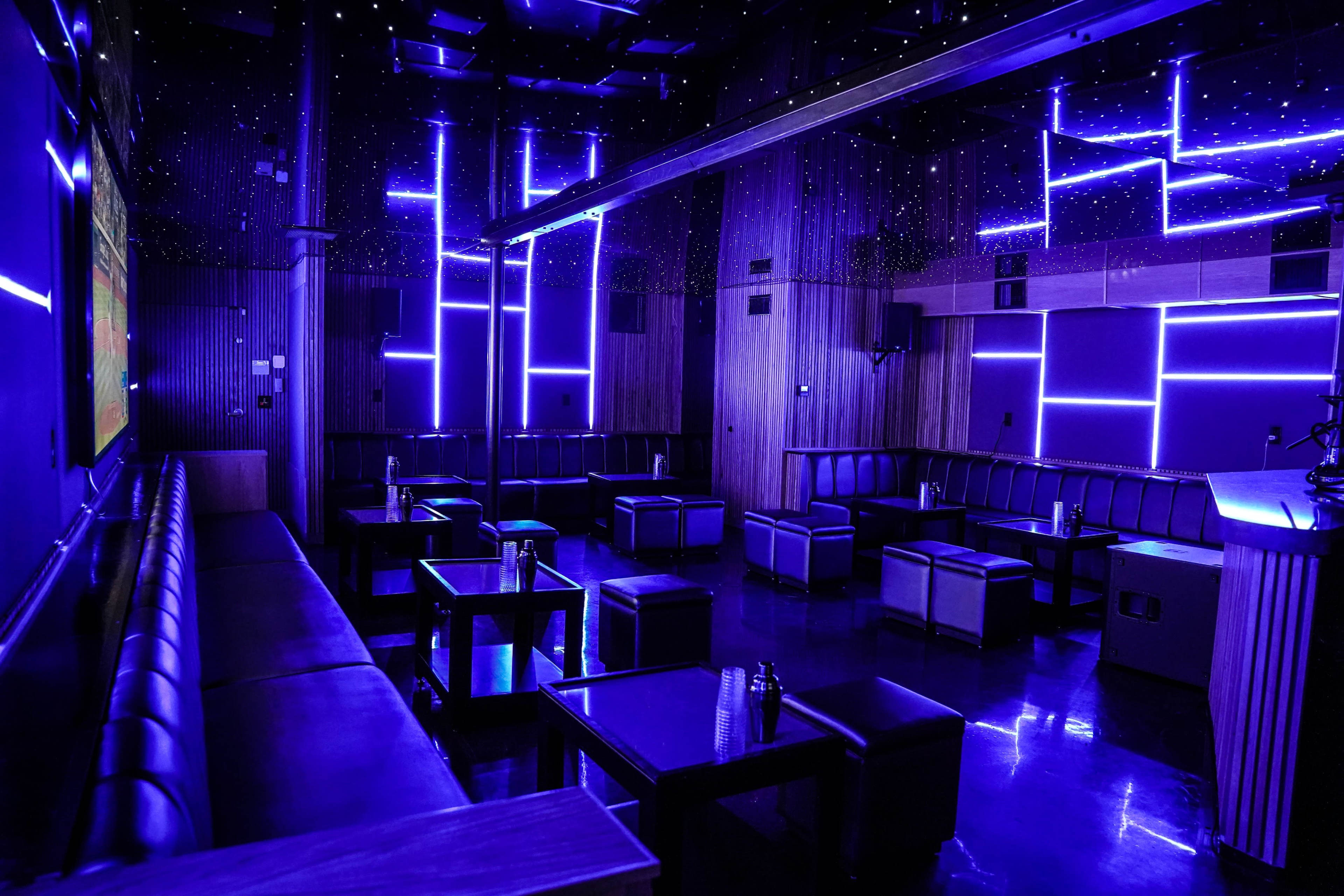 The image shows a dimly lit lounge with purple LED lighting, featuring black leather seating arranged around small tables.