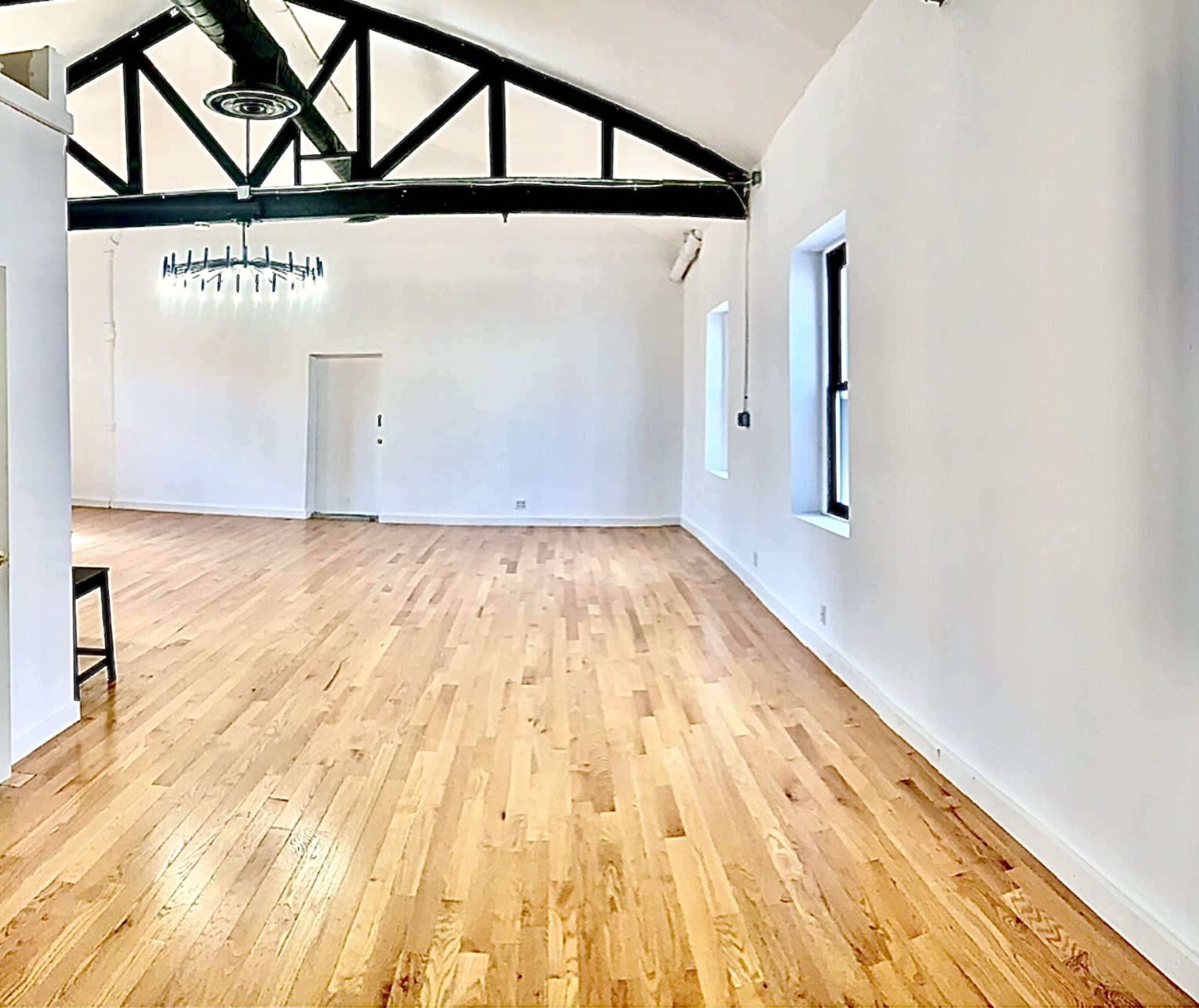 The image shows a spacious, well-lit room with wooden flooring, high ceilings, and a black-beamed structure.