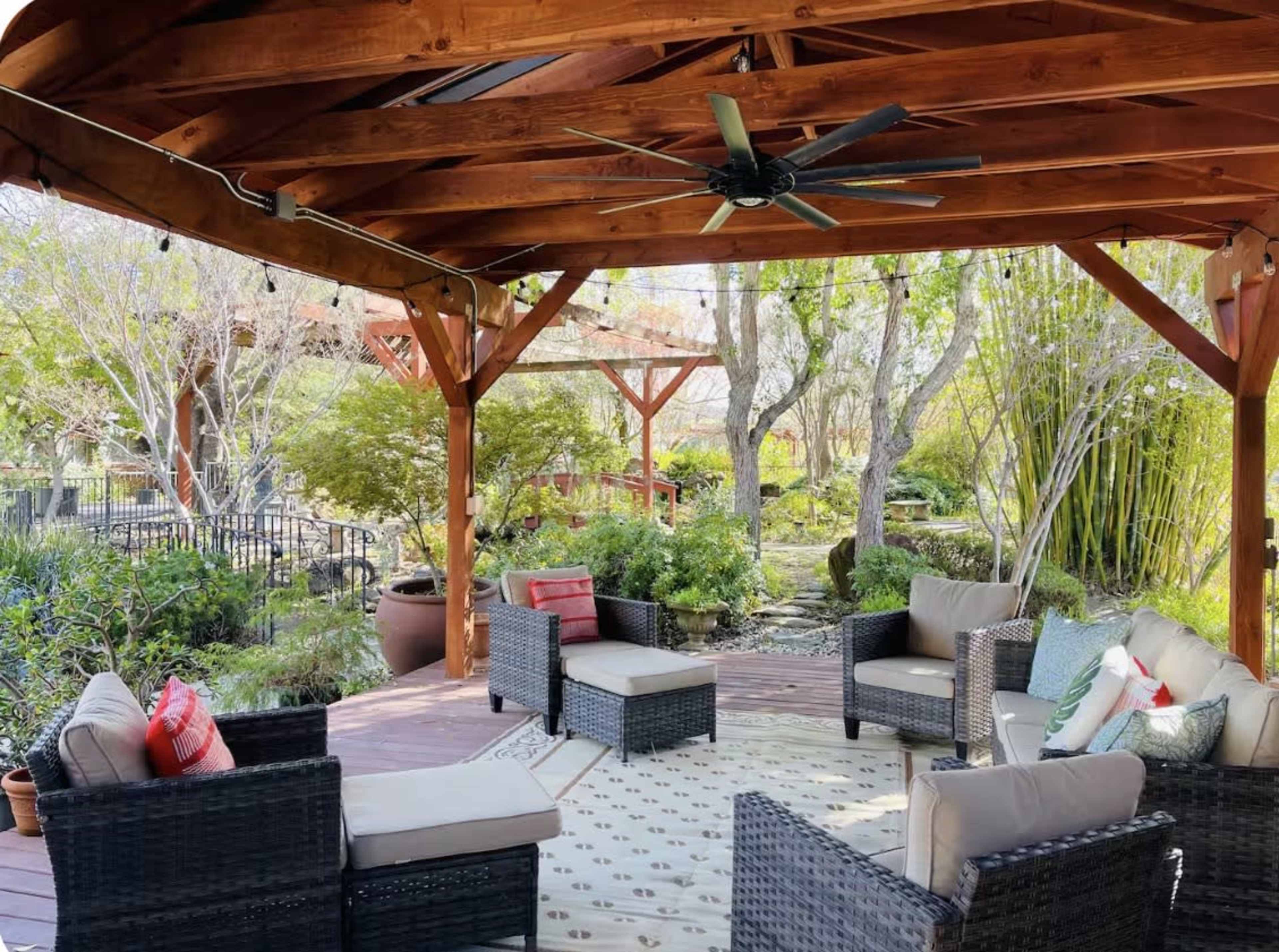 A shaded outdoor seating area features wicker furniture arranged around a patterned rug under a wooden canopy.
