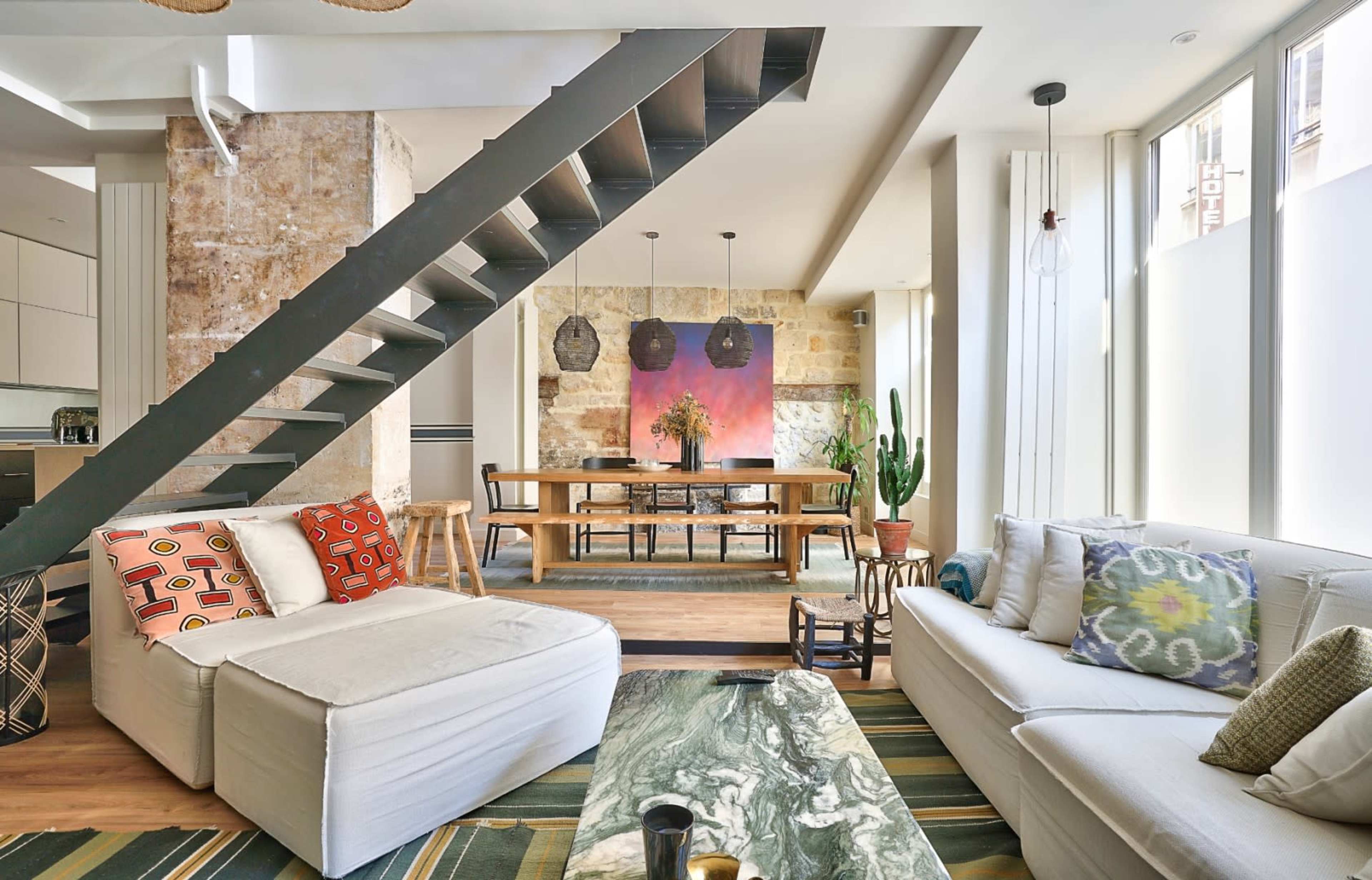A modern living space features a staircase leading to a loft area, with a cozy seating arrangement and a large dining table illuminated by natural light.