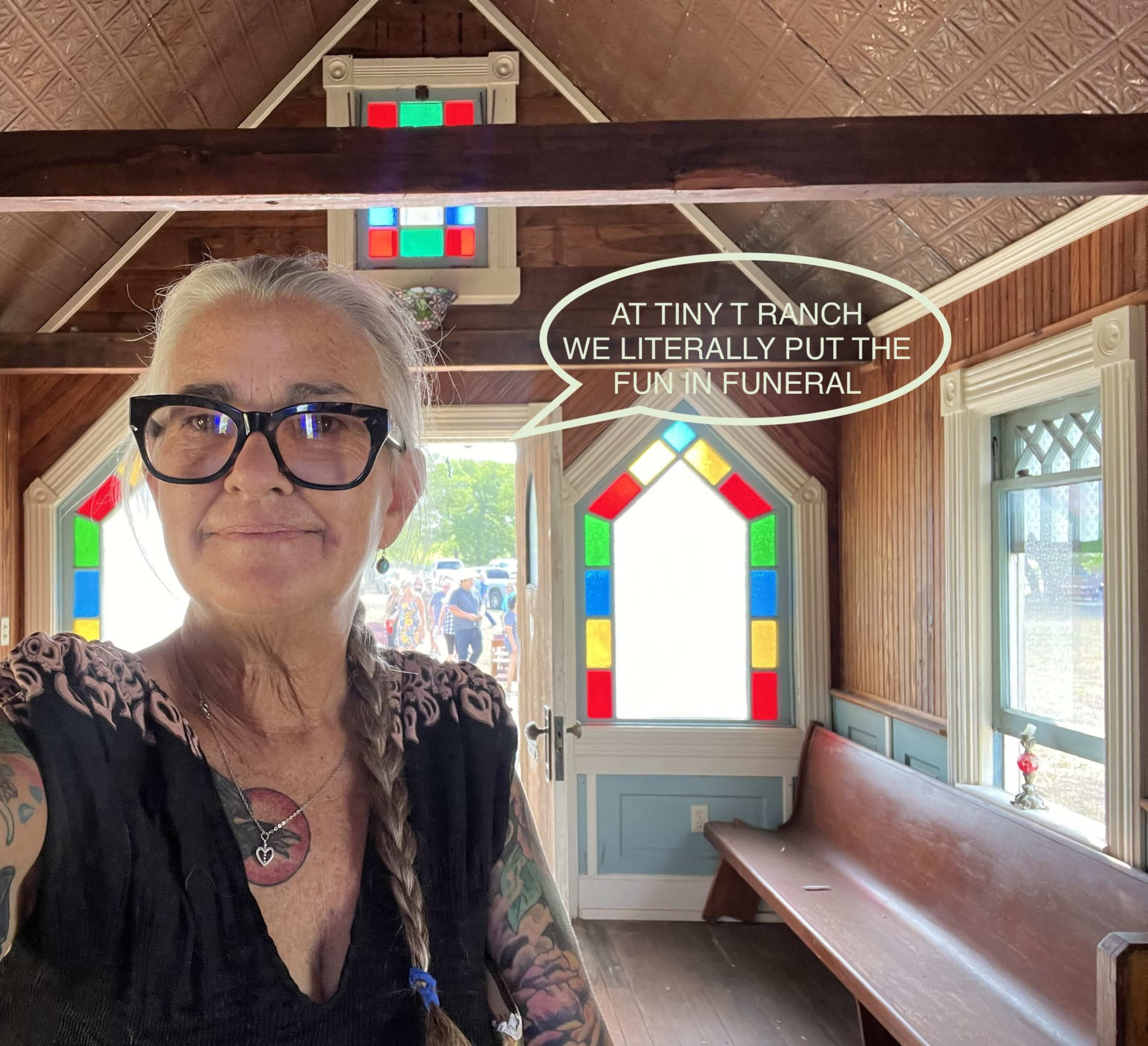 A woman stands inside a small, colorful chapel with stained glass windows, holding a phone and smiling at the camera.
