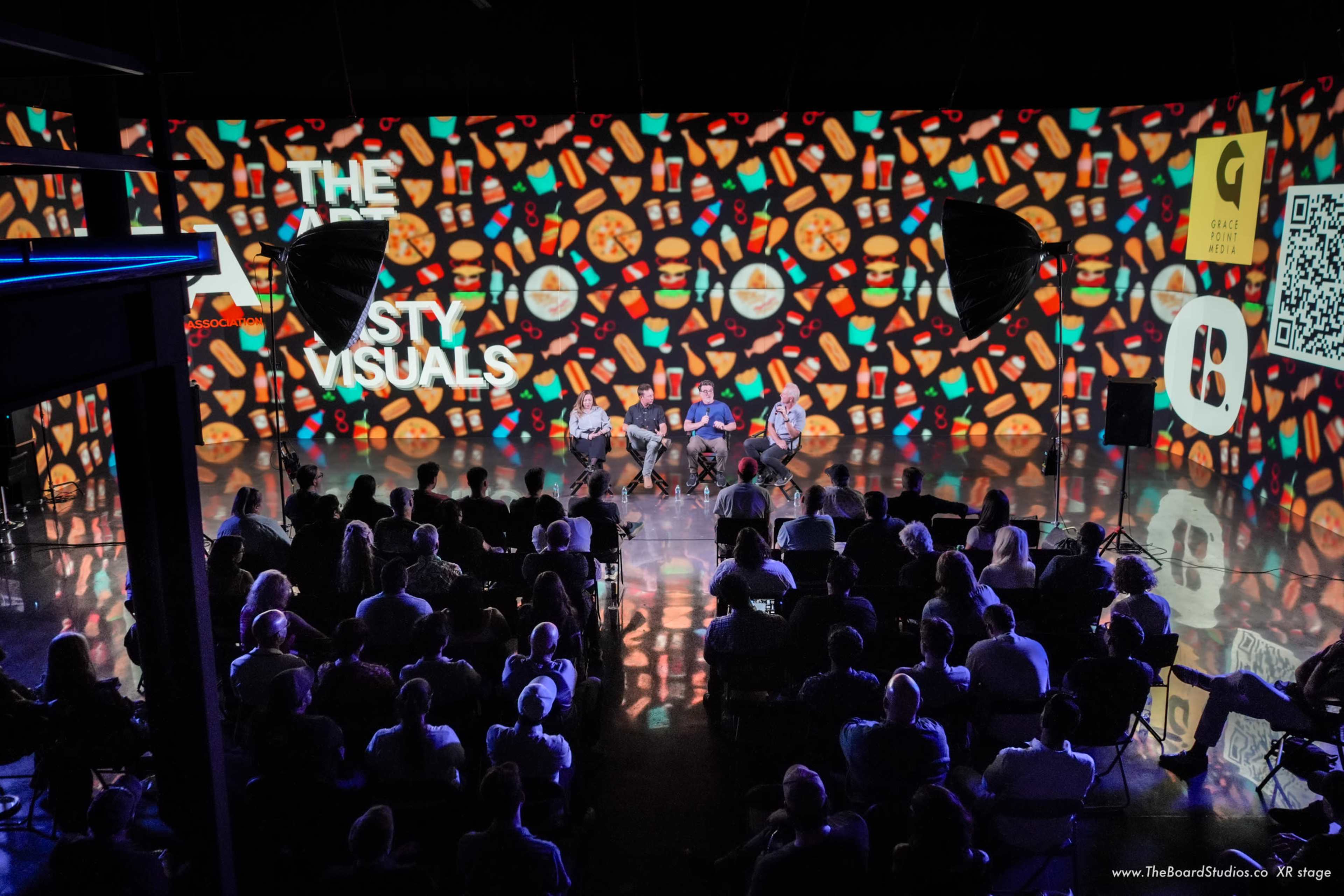 A group of speakers is seated on a stage in front of a large audience, with colorful visuals projected on the backdrop.