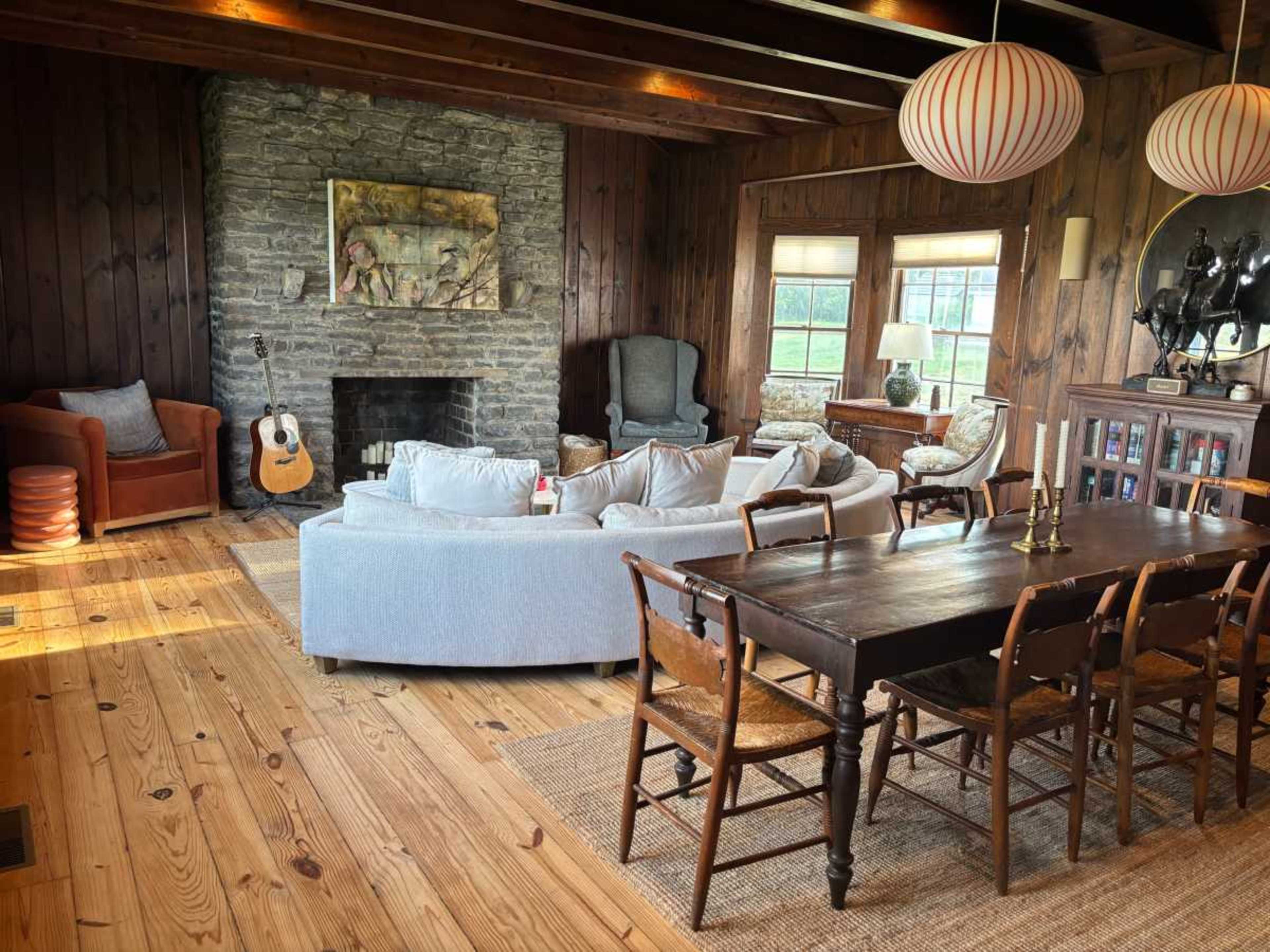 The image shows a rustic living and dining area with wooden walls, a stone fireplace, a mix of seating options, and a long dining table.
