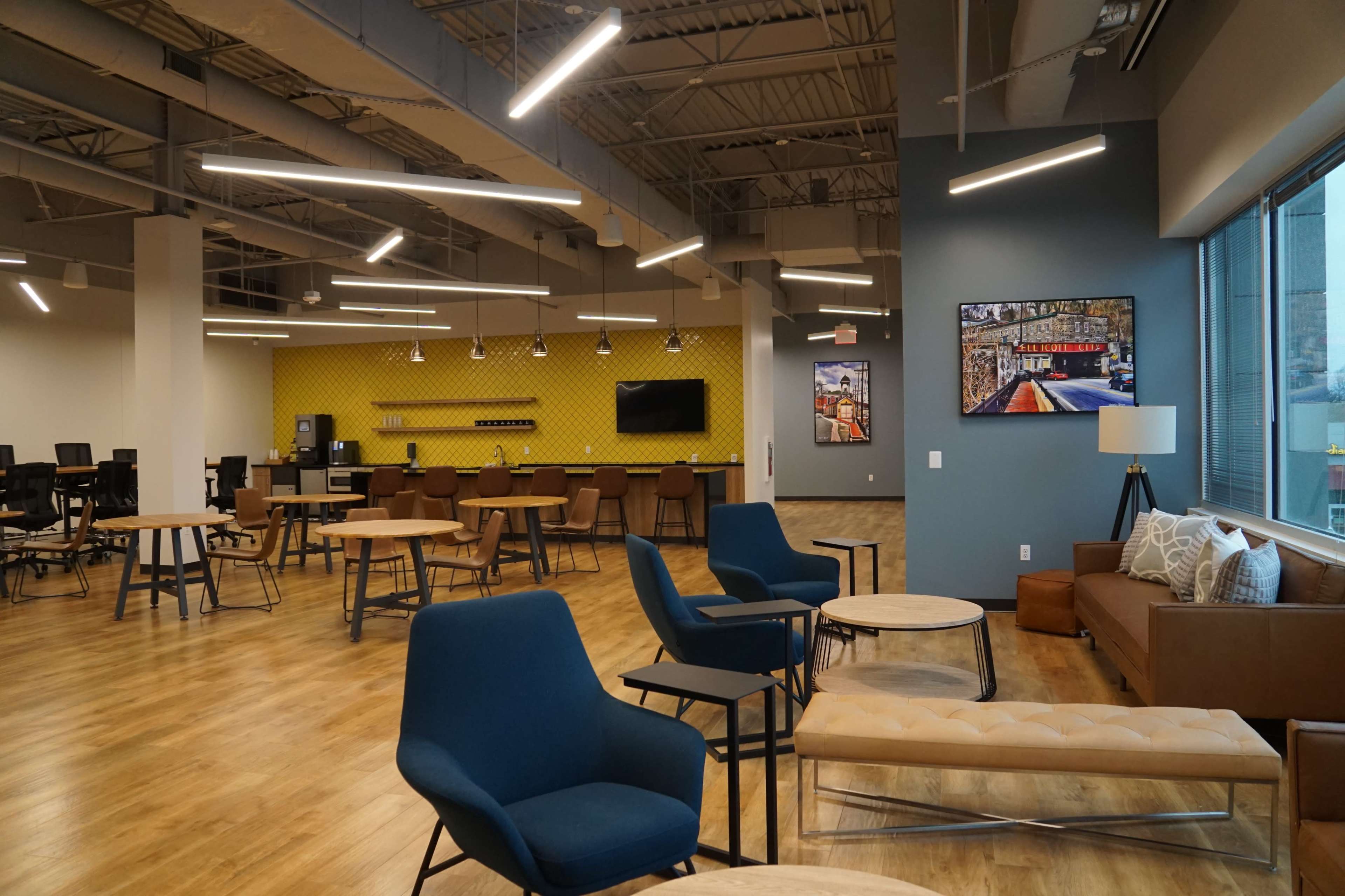 The image shows a spacious, modern office interior featuring wooden flooring, various seating areas, and a vibrant yellow accent wall.