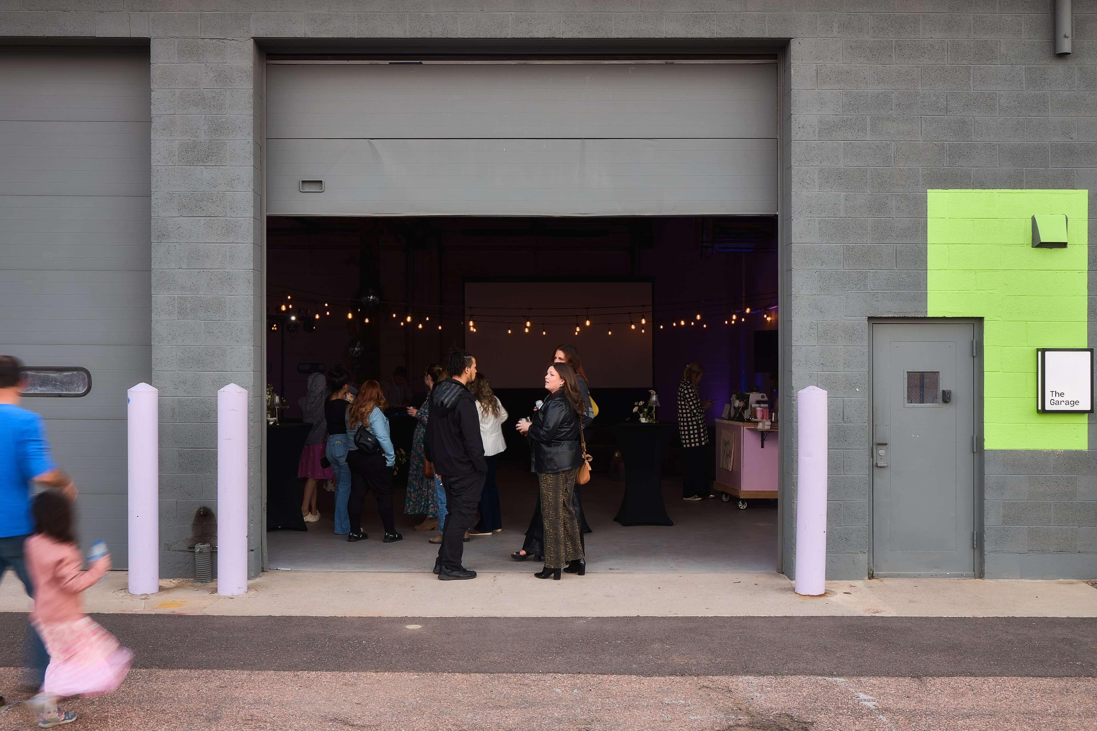 A group of people gather inside a brightly lit room seen through a garage-style opening, with some individuals engaged in conversation while others browse the space.