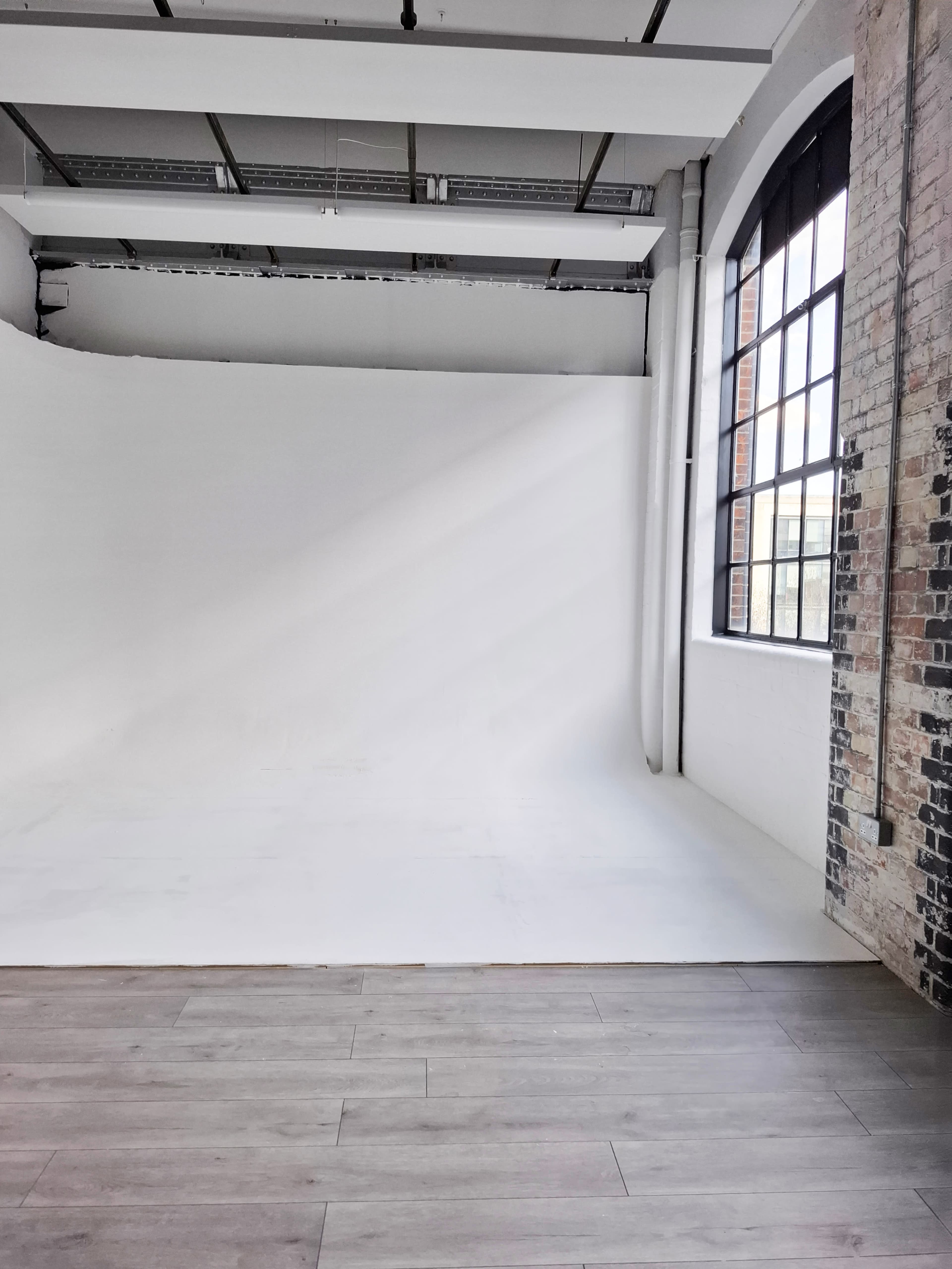 The image shows a spacious photography studio featuring a white backdrop, large windows with natural light, and wooden flooring.