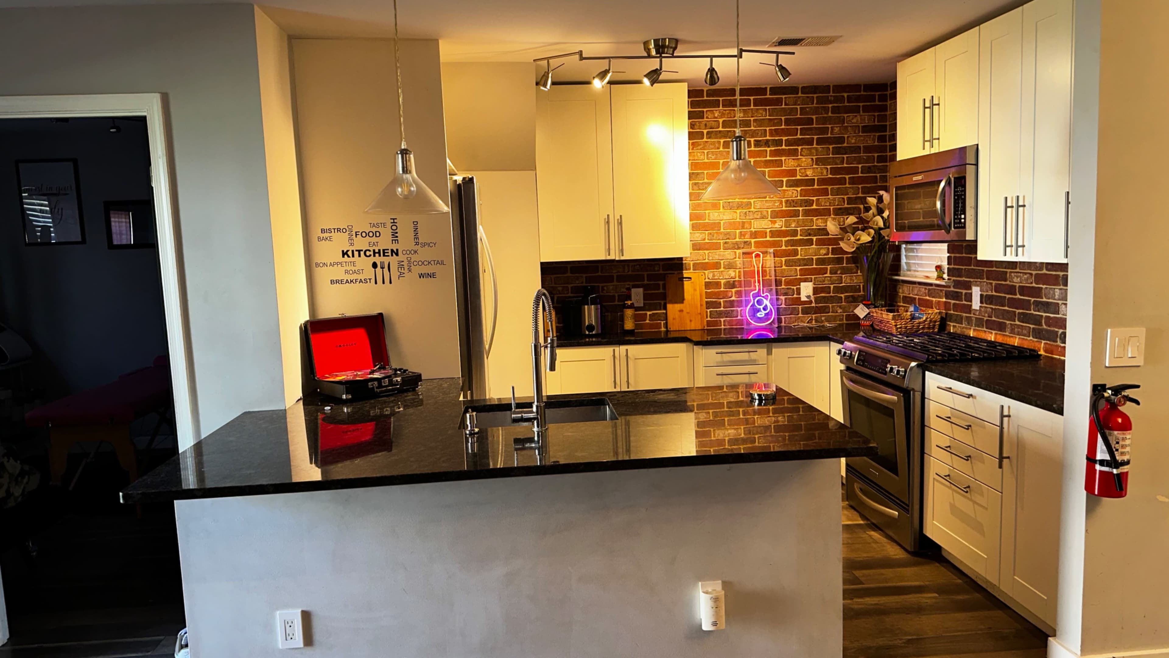 A modern kitchen with white cabinets, a stainless steel refrigerator, and a black granite countertop, featuring a brick accent wall and under-cabinet lighting.