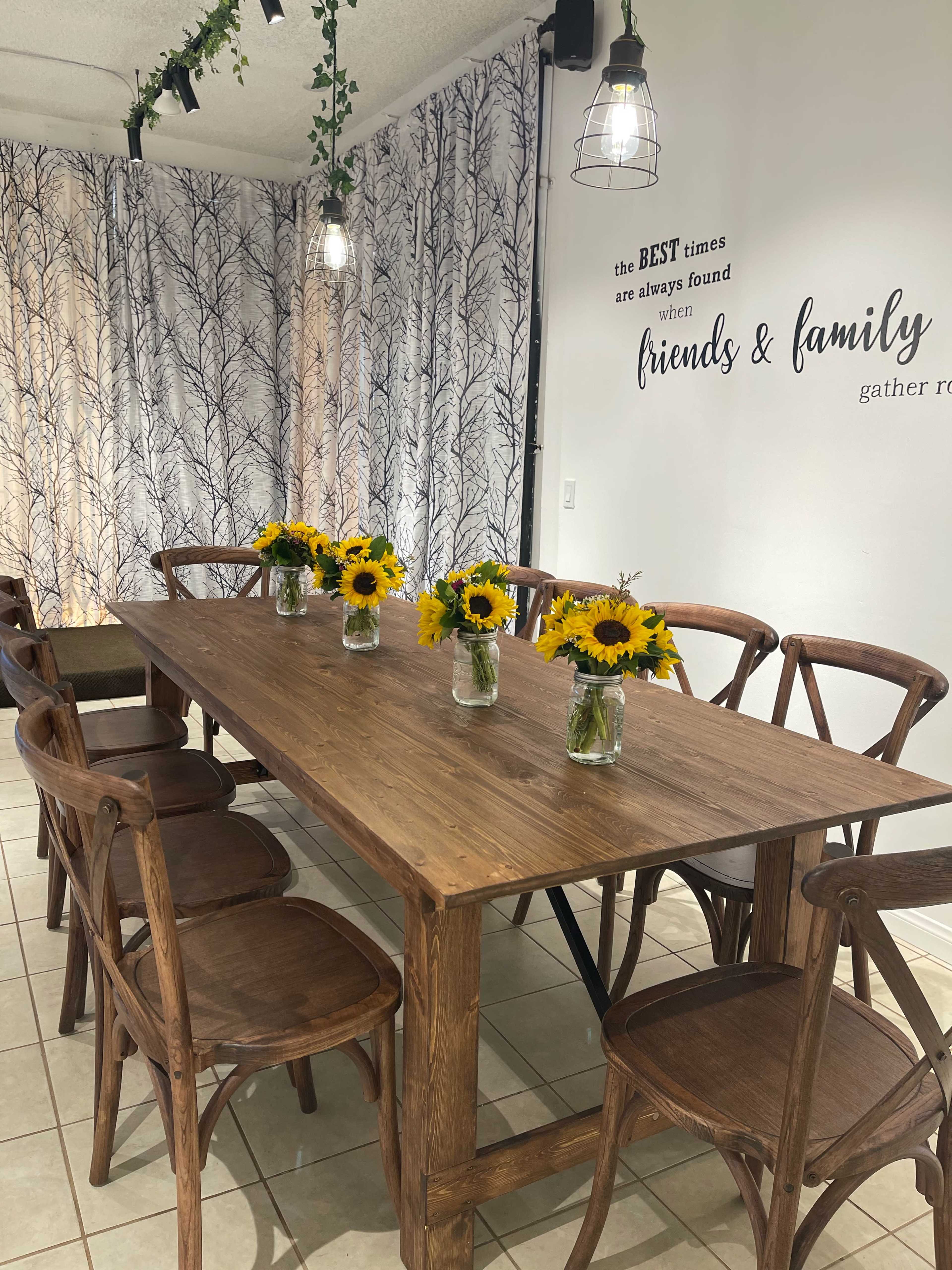 A long wooden dining table is set with mason jars of sunflowers, surrounded by wooden chairs, in a room decorated with a tree-patterned curtain and a wall inscription about friends and family.