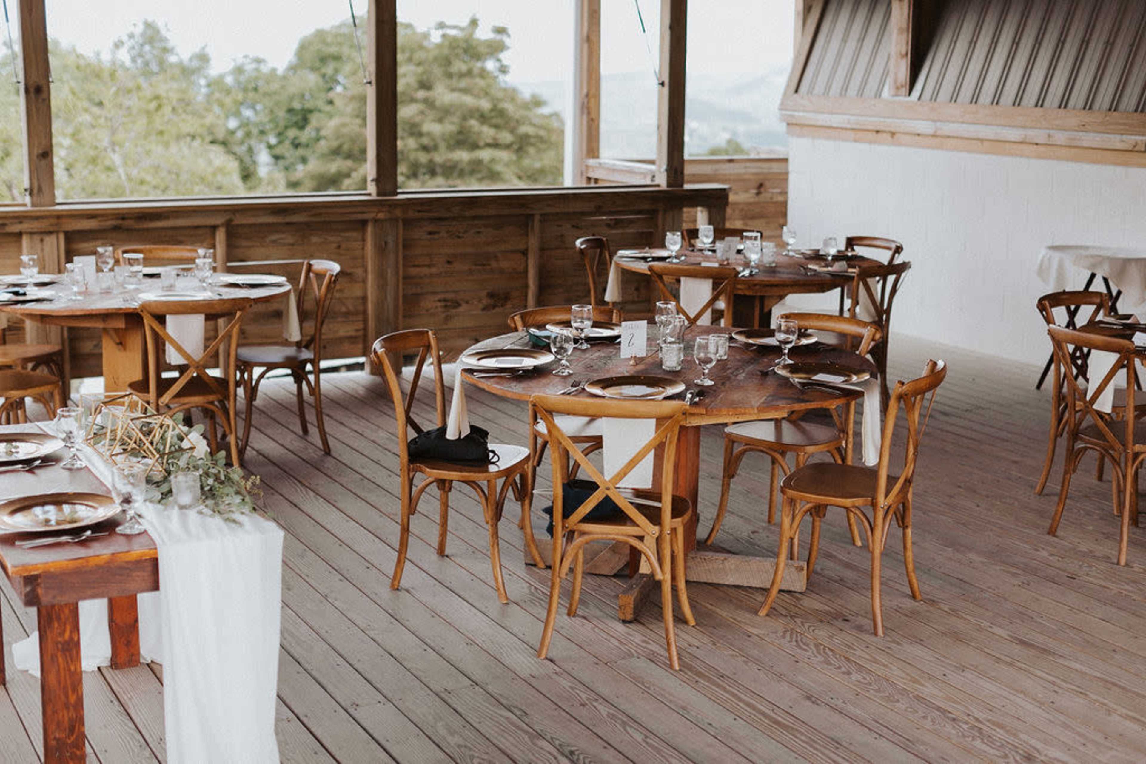 The scene features an elegantly arranged dining area with wooden tables and chairs, set up for guests, overlooking a natural landscape.