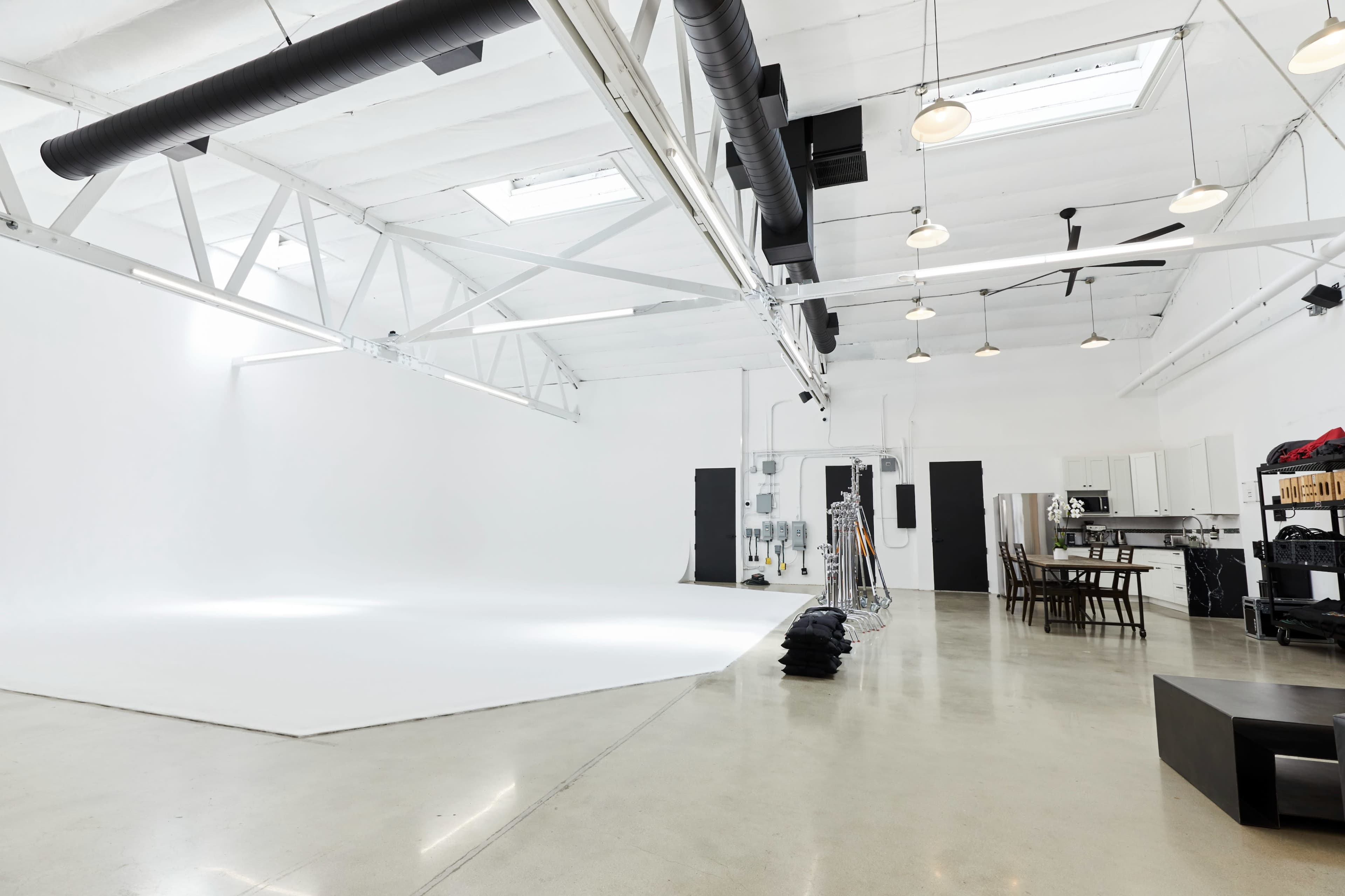 The image shows a spacious, minimalist studio with a white backdrop, polished concrete floors, and a kitchen area in the background.