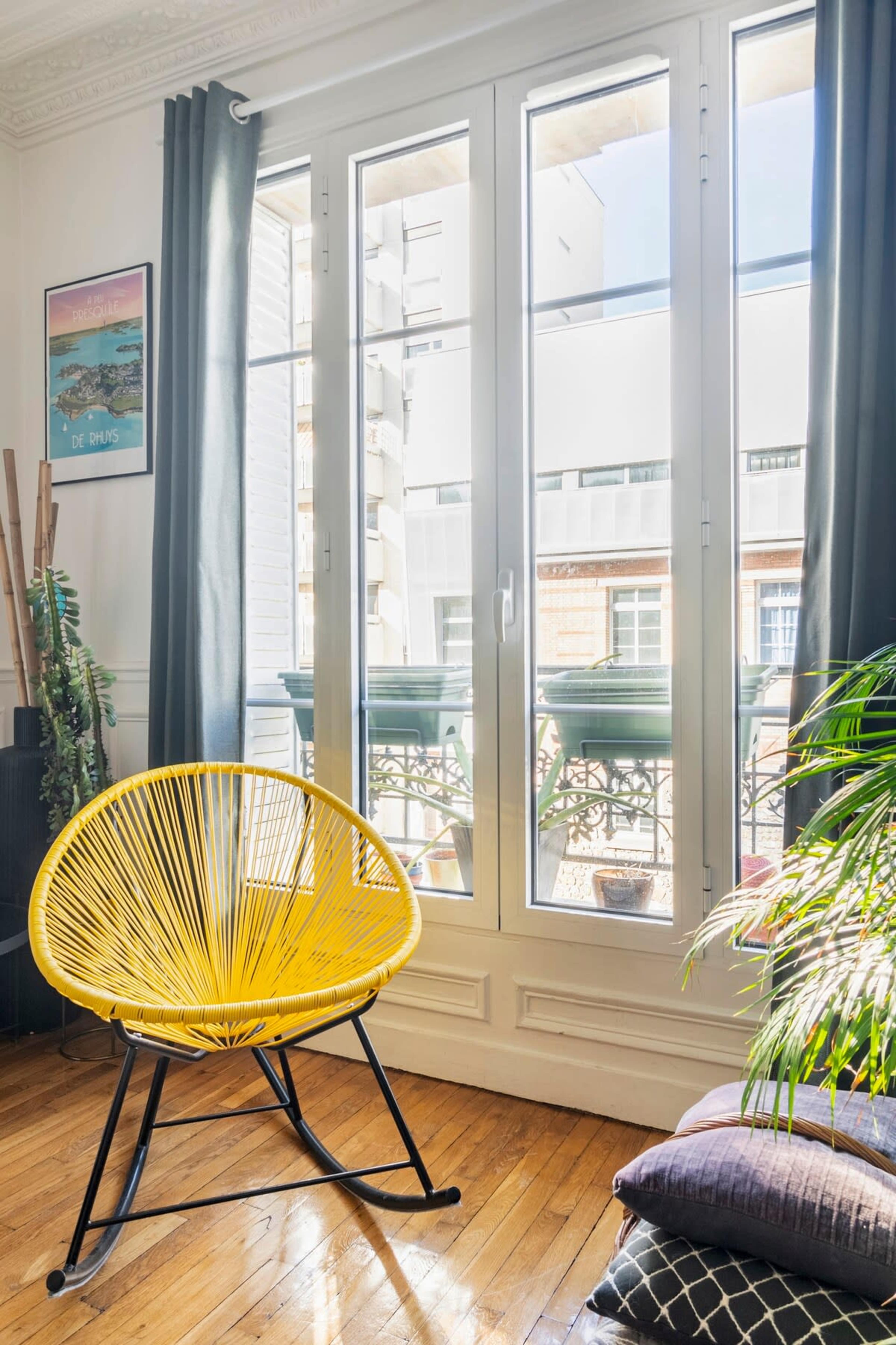 A yellow chair is positioned near large windows that open onto a balcony, with sunlight illuminating the room.