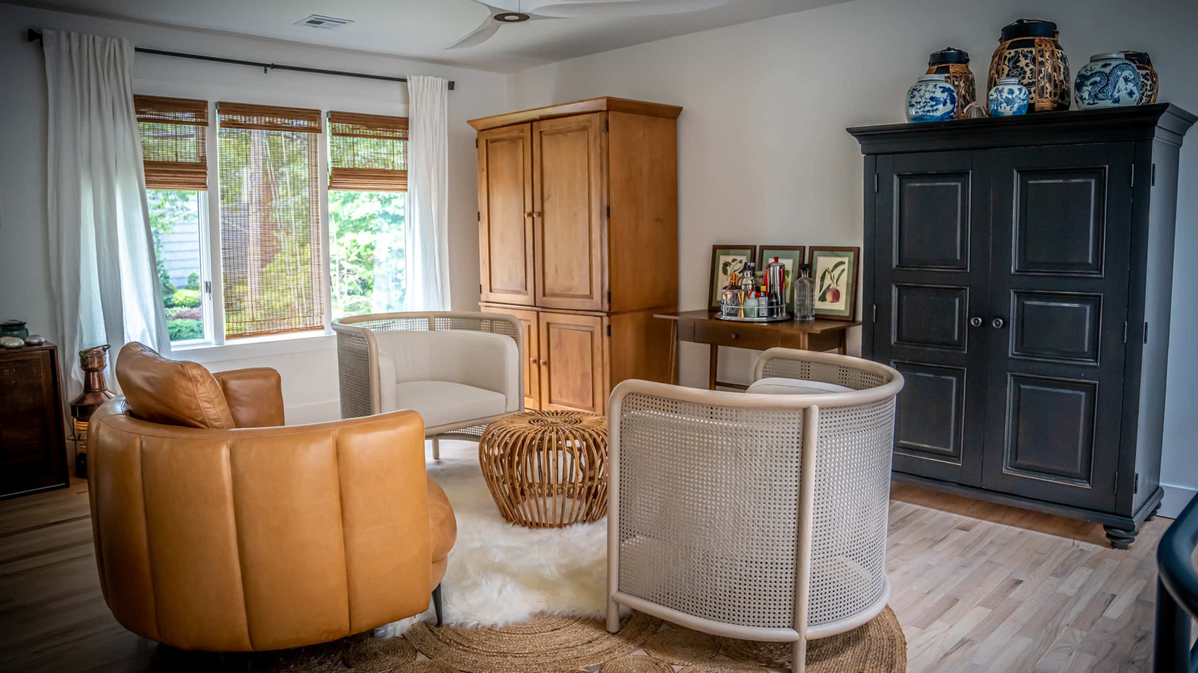 A cozy living room featuring two circular chairs, a brown leather armchair, a wooden cabinet, and a decorative woven table, illuminated by natural light from a window.