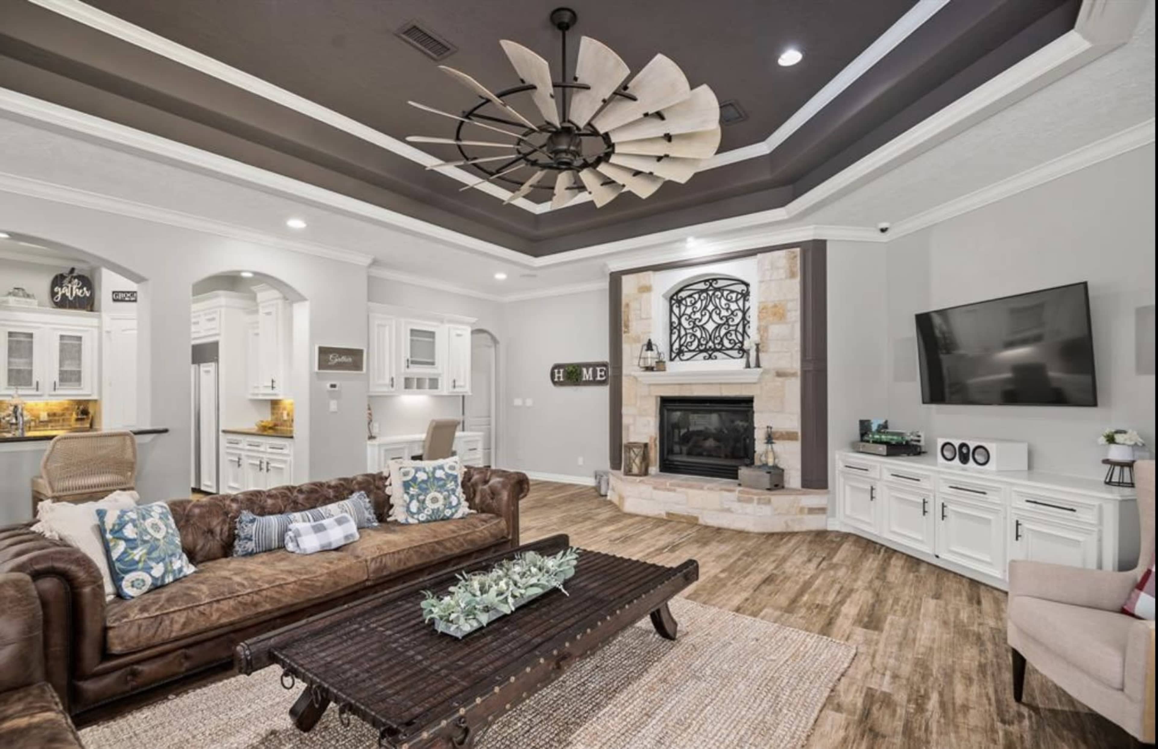 A living room featuring a brown leather sofa, a wooden coffee table, a stone fireplace with a television above it, and a ceiling fan.