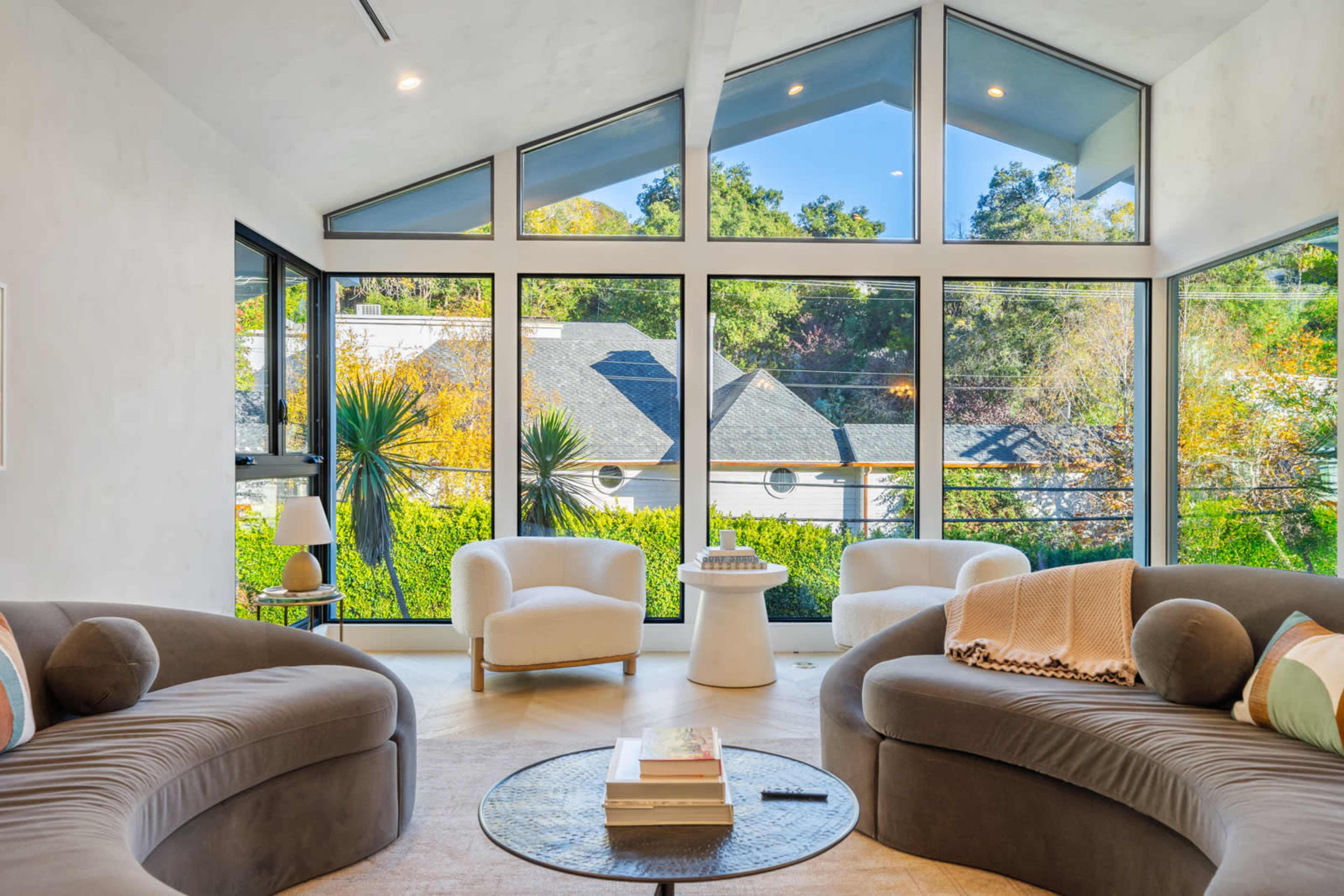 The image shows a modern living room with large windows offering a view of greenery and a nearby house, featuring curved sofas and a round coffee table.