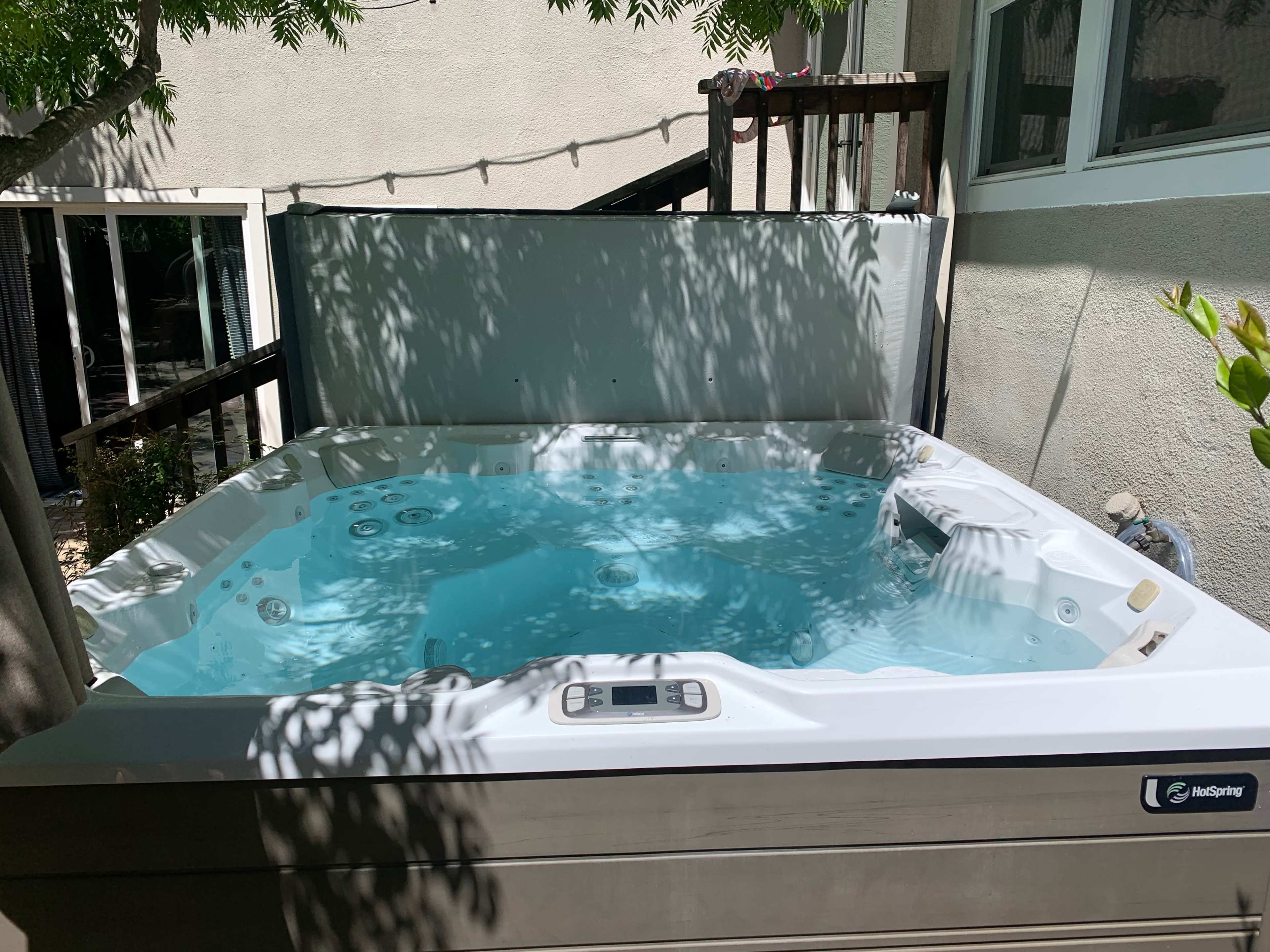 A hot tub filled with clear water is situated in a shaded area surrounded by a fence and trees.