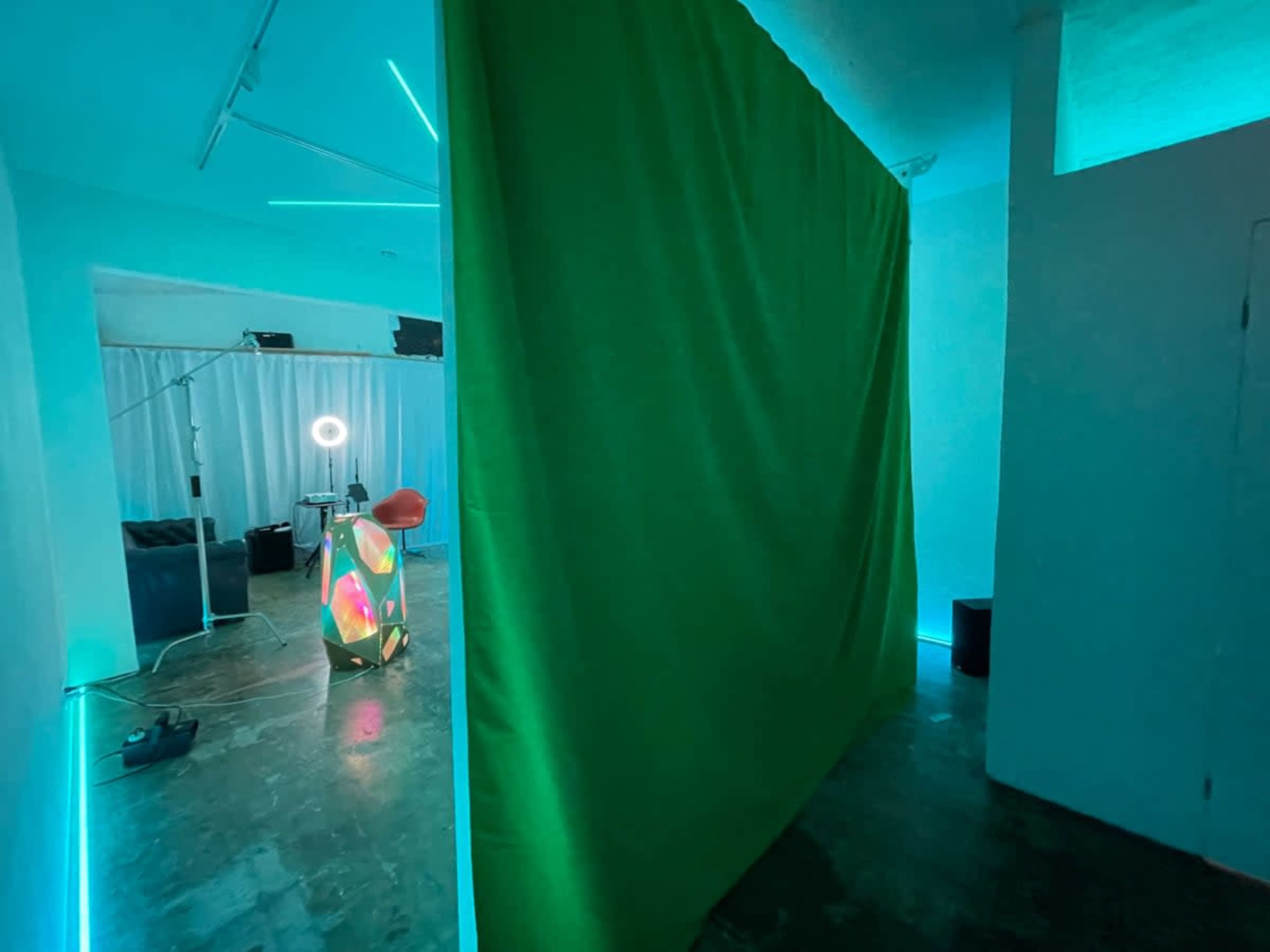 The image shows a room with a green curtain blocking a part of the space, illuminated by blue and green lights, with various equipment and furniture visible in the background.