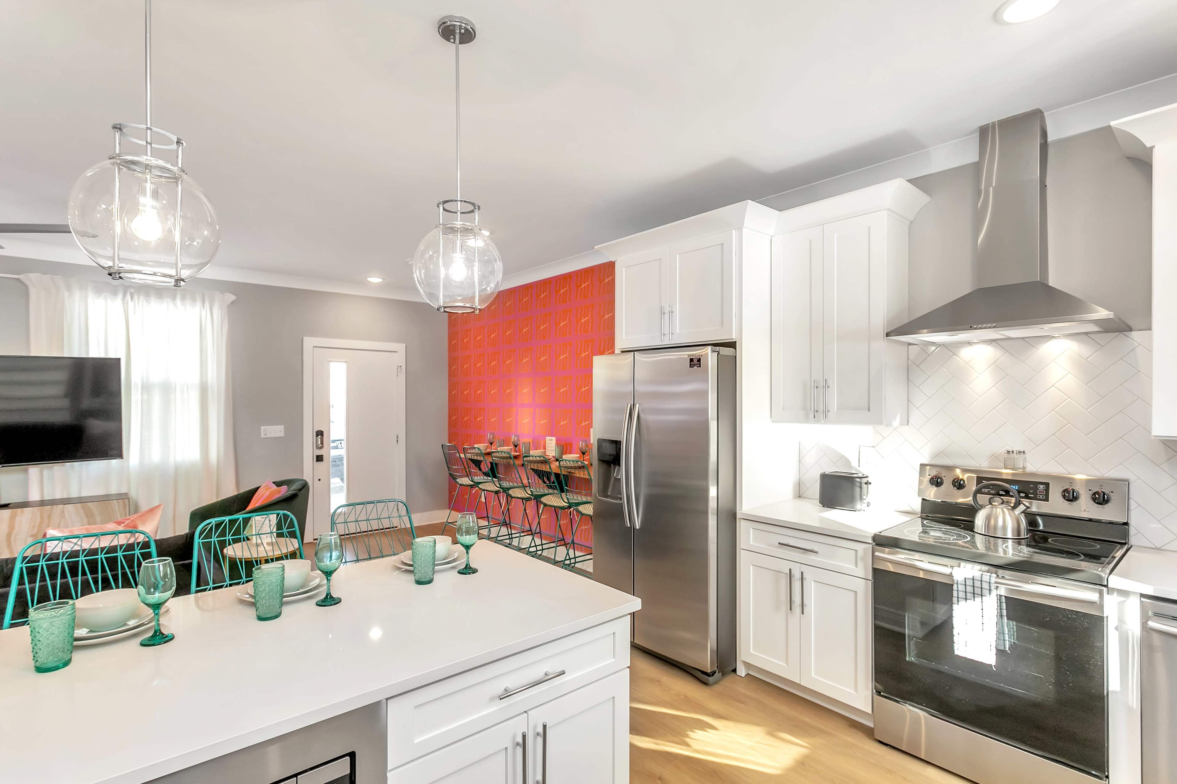 A modern kitchen features white cabinetry, stainless steel appliances, and a vibrant orange accent wall with a dining area visible in the background.