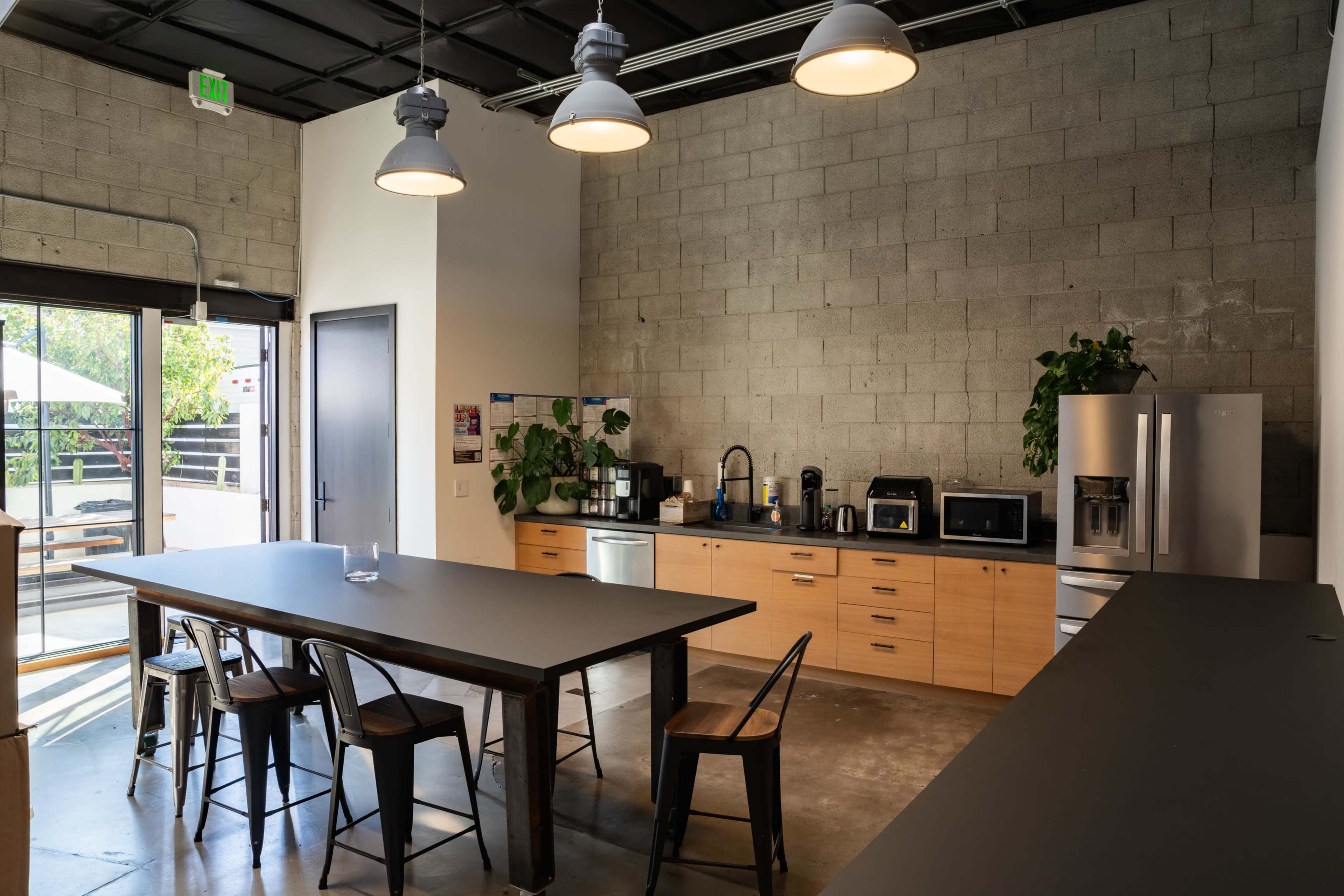 The image shows a modern kitchen area with a large table, various appliances, and plants, alongside a glass door that leads to an outdoor space.